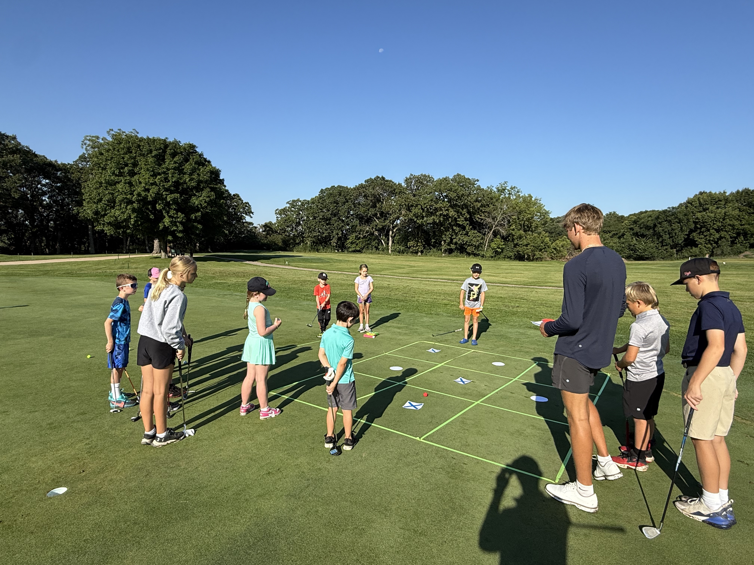 Children and an adult gathered on a golf course, playing a golf game on the grass under a clear blue sky with trees in the background.