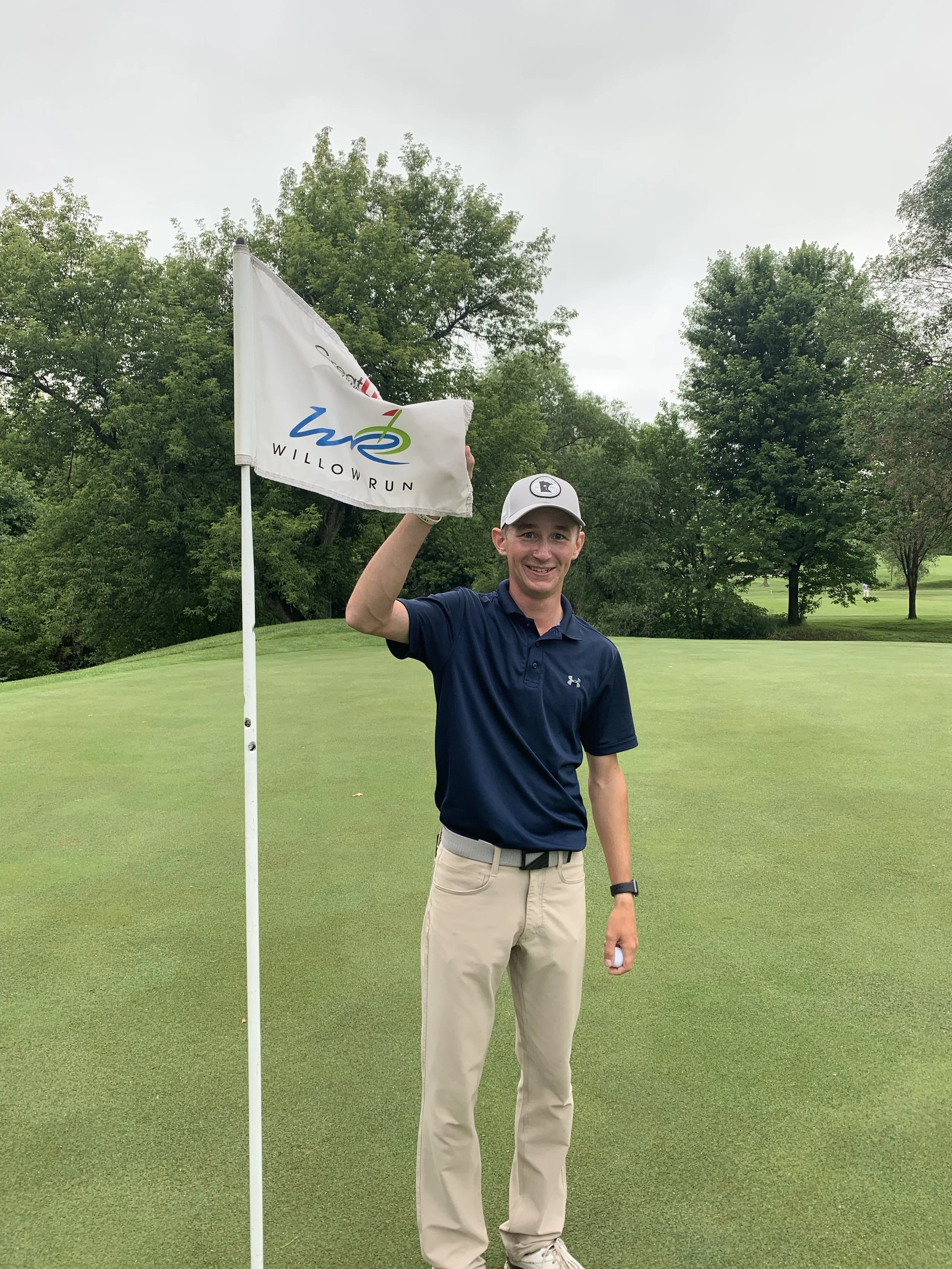 A young man in khaki pants and a navy polo shirt is standing on a golf green, holding a Willow Run golf flag. He is smiling and wearing a white cap and a black watch, with trees and a cloudy sky in the background.