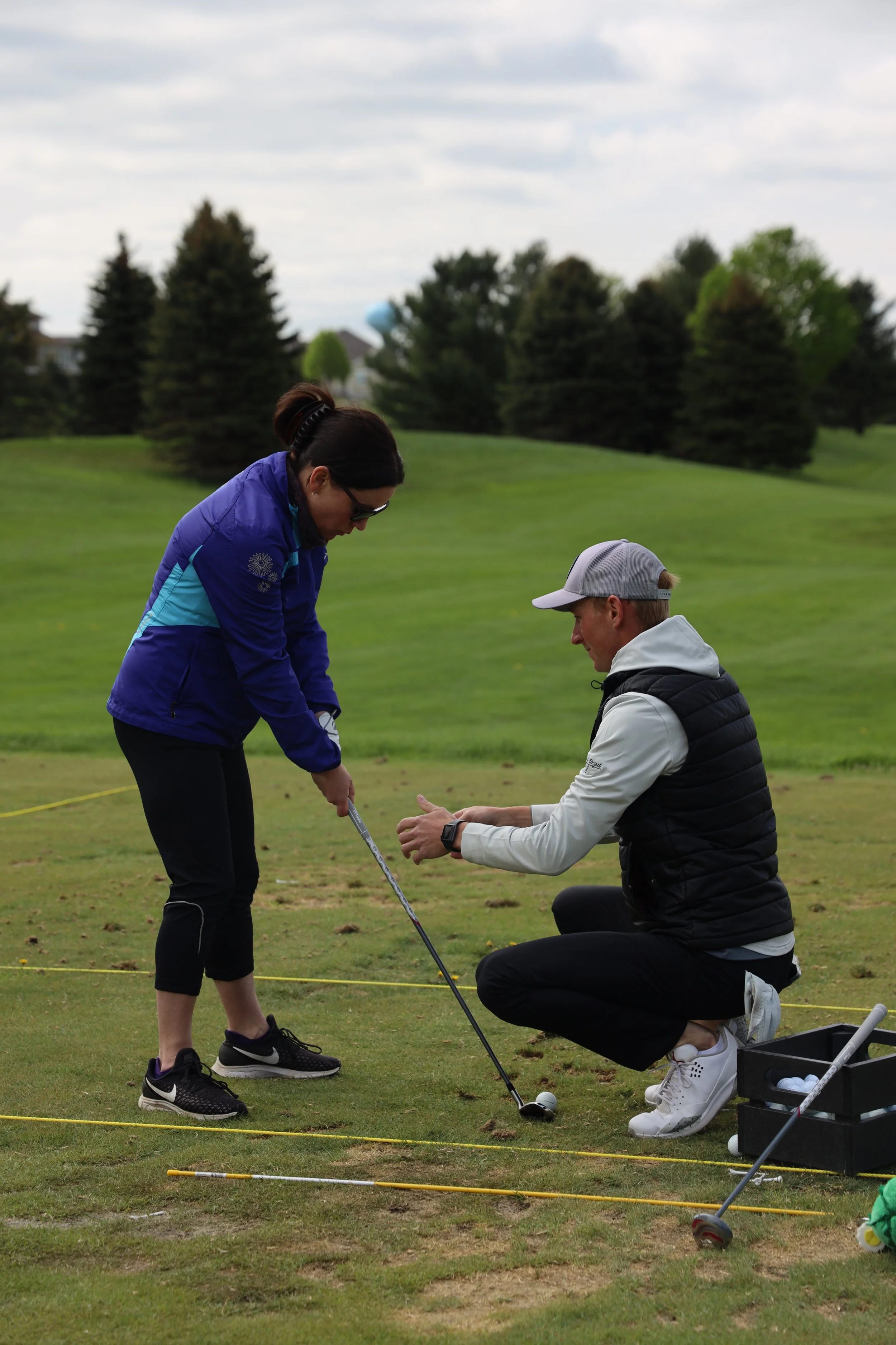 A woman learning golf from a man on a golf course, with trees and a cloudy sky in the background.
