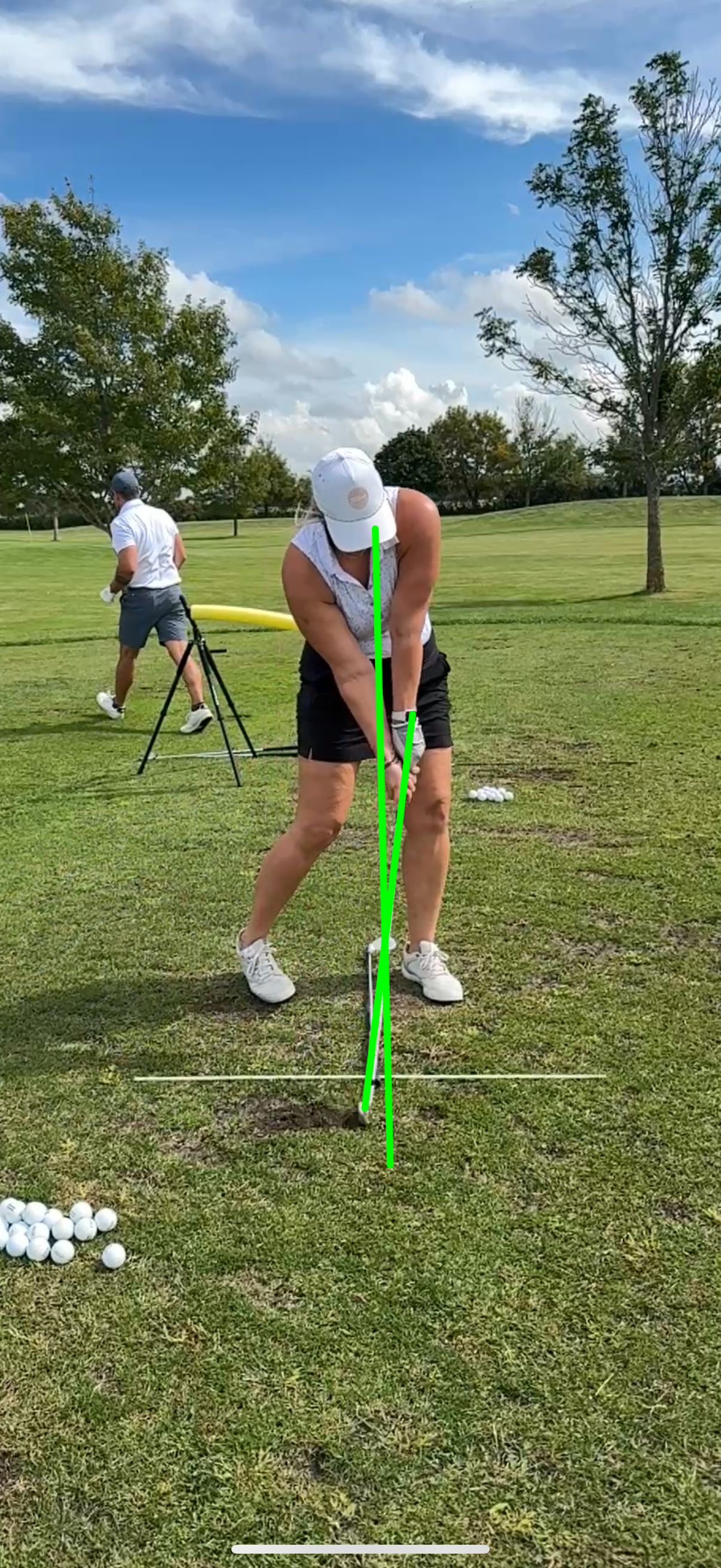 A woman in a white cap and black shorts practicing golf swing on a grassy field, with a male in the background nearby. There are golf balls on the ground and some equipment in the background, under a blue sky with clouds.