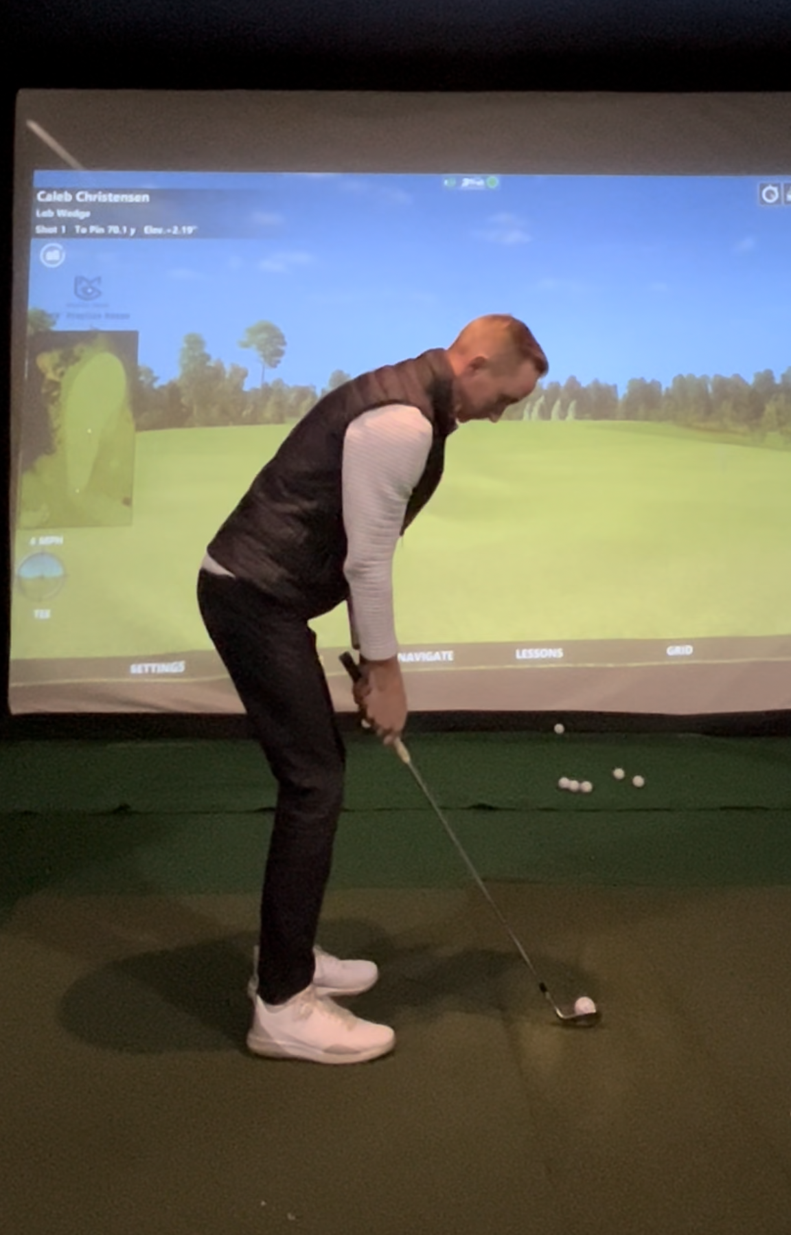 Man practicing indoor golf in front of a digital golf simulator screen, holding a golf club, with golf balls on the floor.