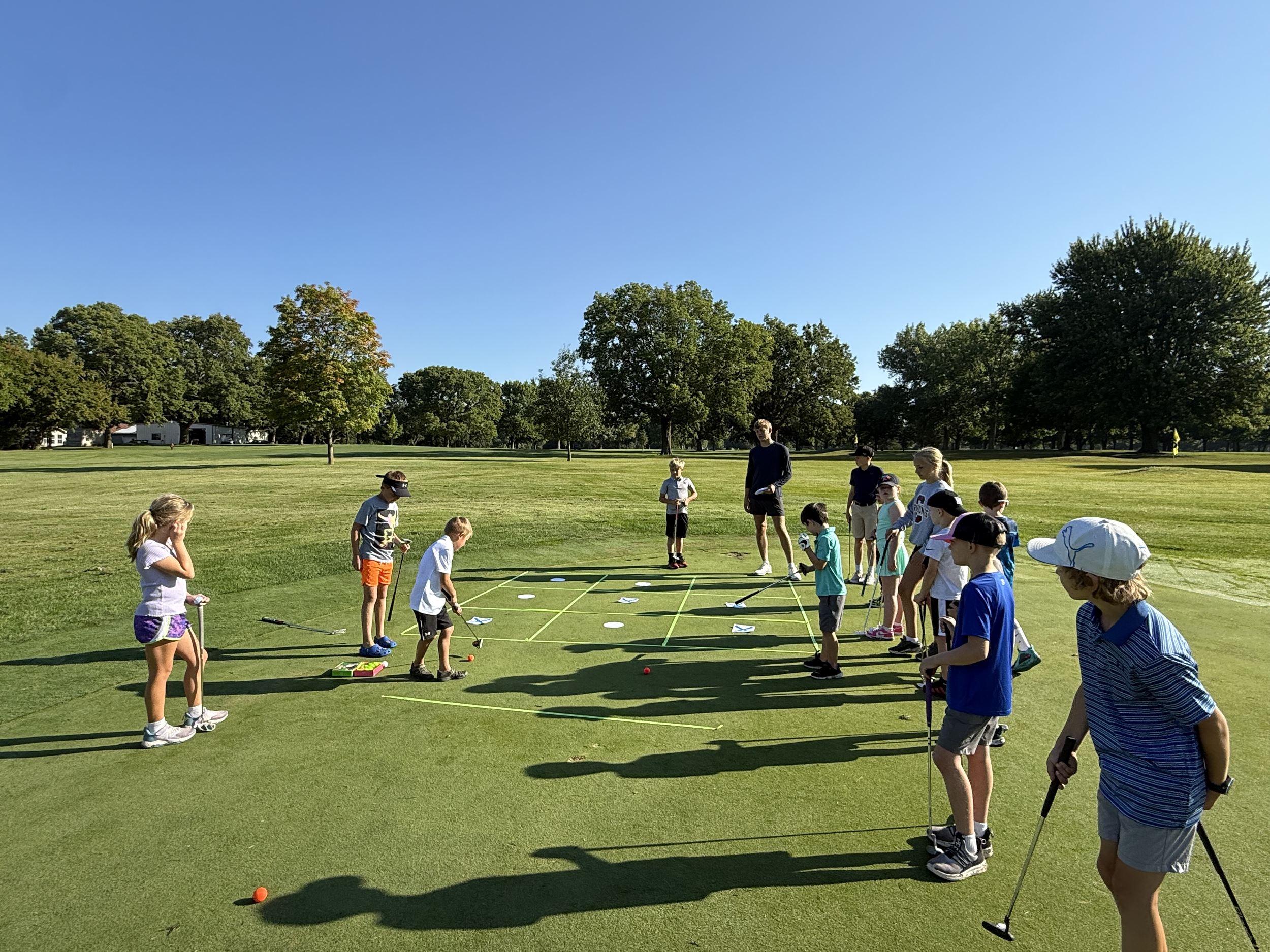 Children and adults practicing golf on a golf course during daytime with a clear blue sky and trees in the background.