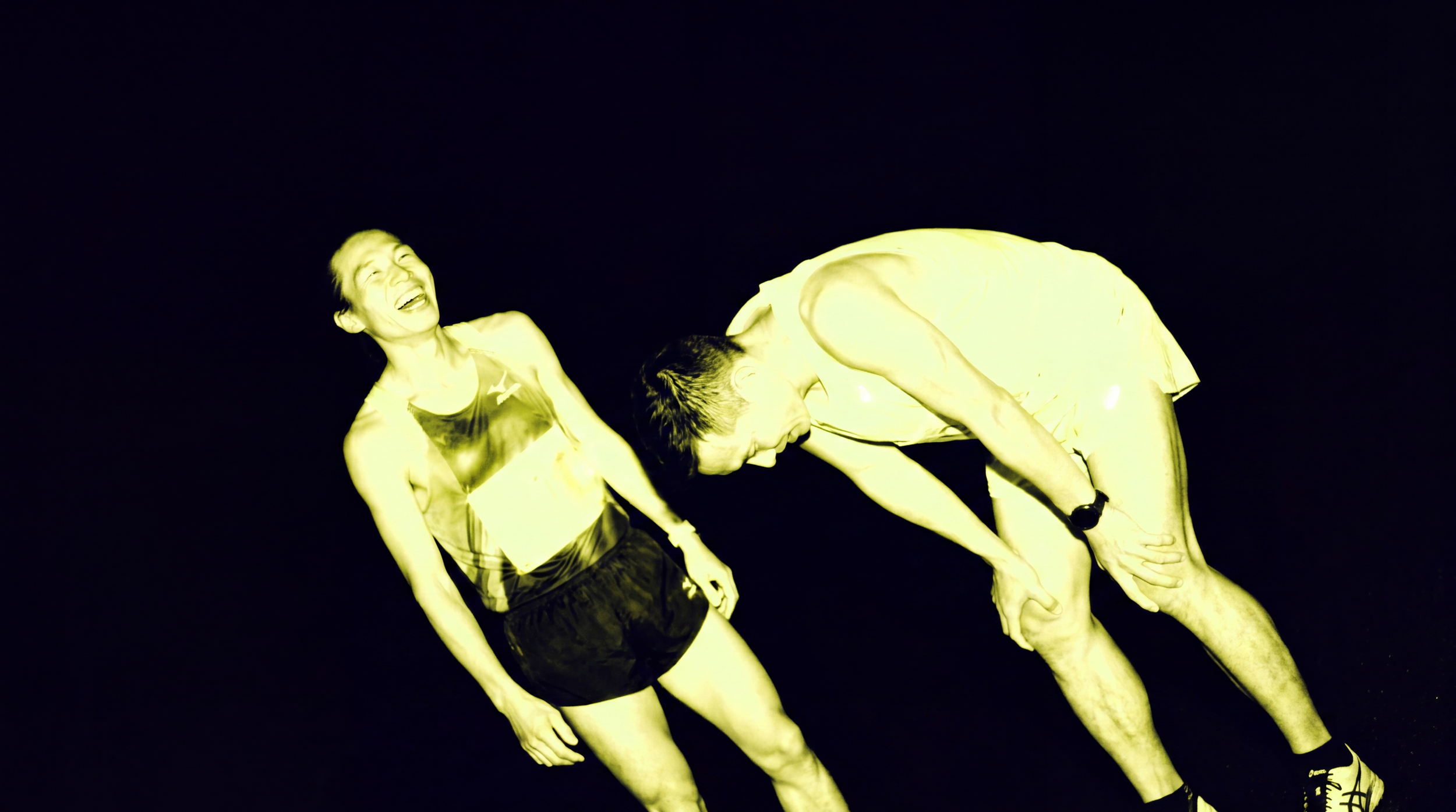 Two people, a woman and a man, are laughing and bowing at night. The woman is wearing a sleeveless top, and the man is wearing a short-sleeved shirt and shorts. The background is dark, and the photo has a yellow tint.