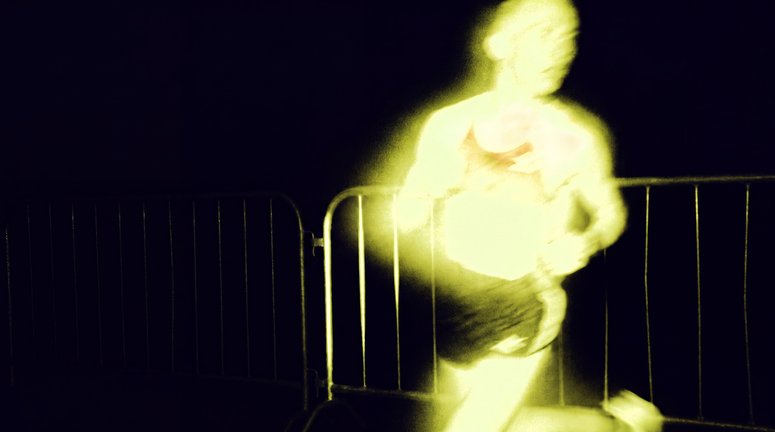 Person standing behind a metal fence, illuminated by a bright yellow light, in a dark environment.