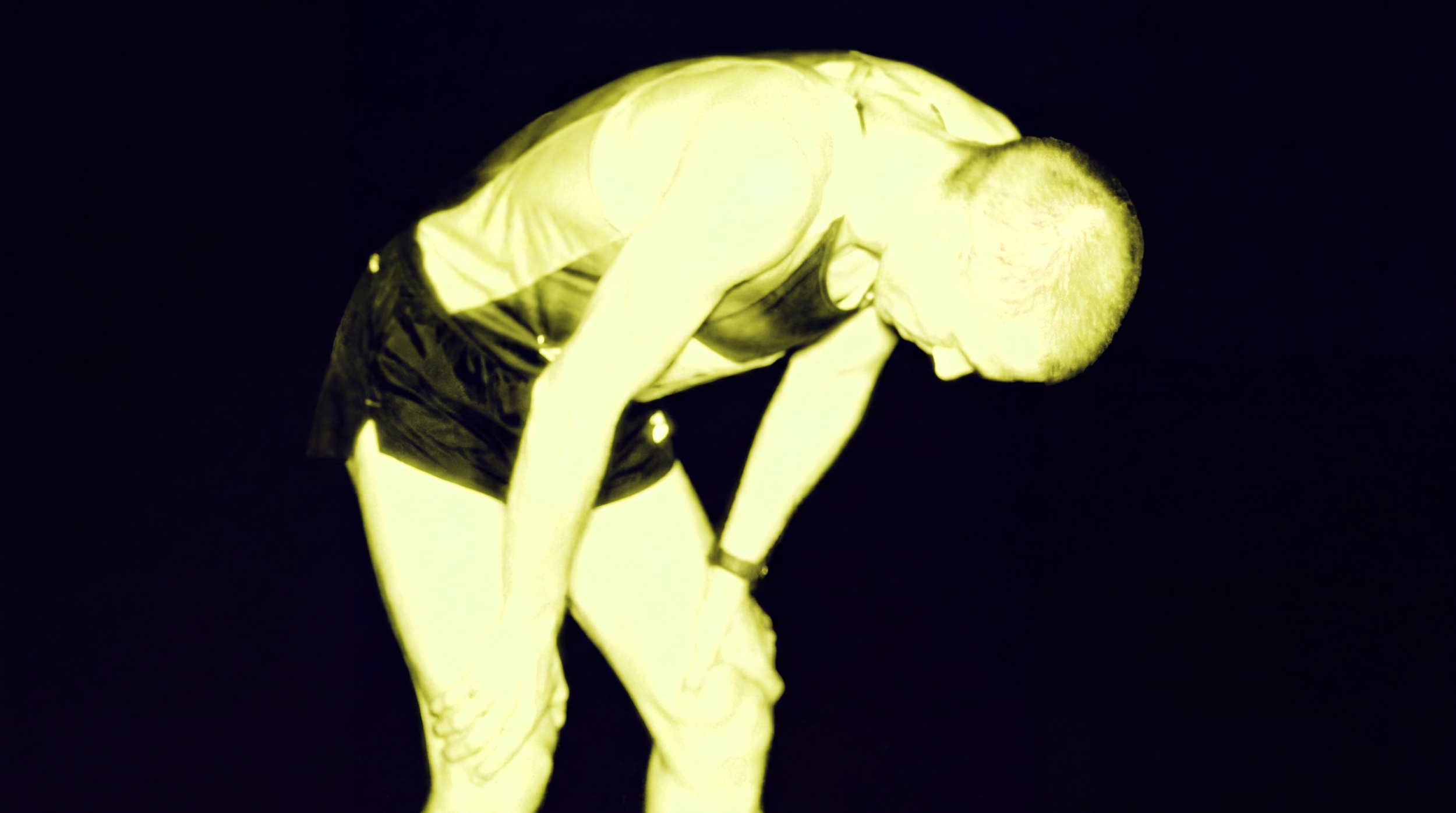 A person with a shaved head wearing a dark shirt, bent over in a dark environment with yellow-tinted lighting.