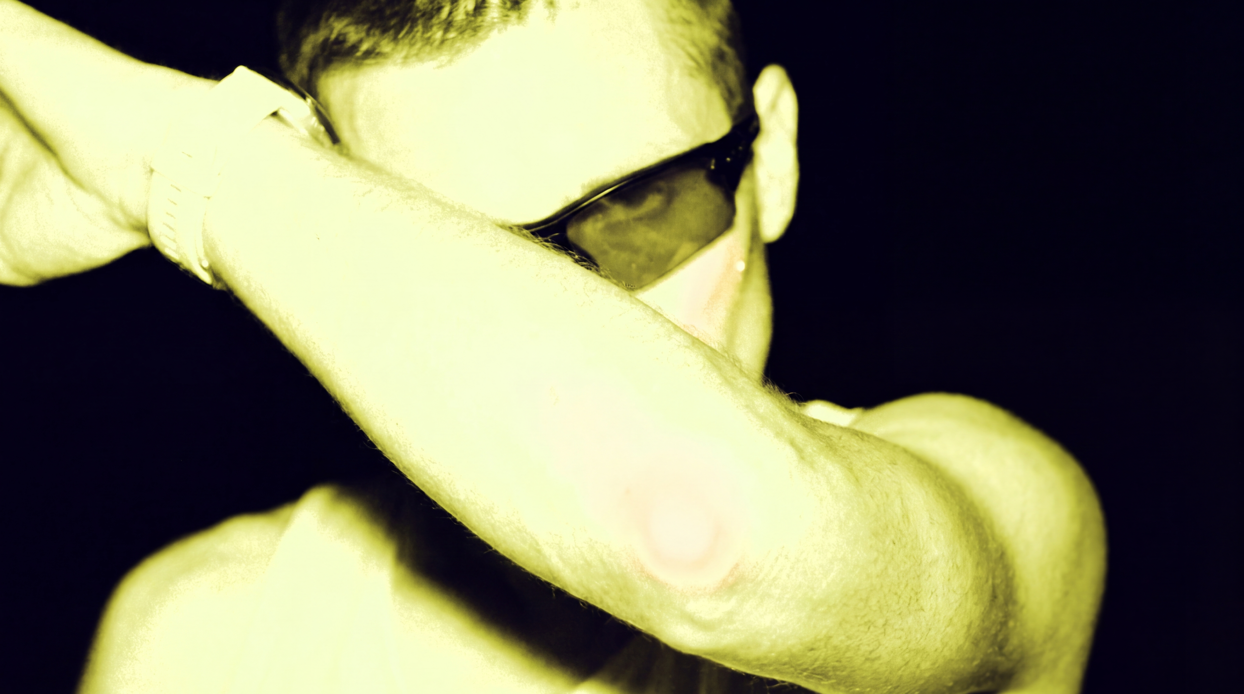 Close-up of a person wearing sunglasses and covering part of their face with their arm, in a high-contrast, yellowish tone.