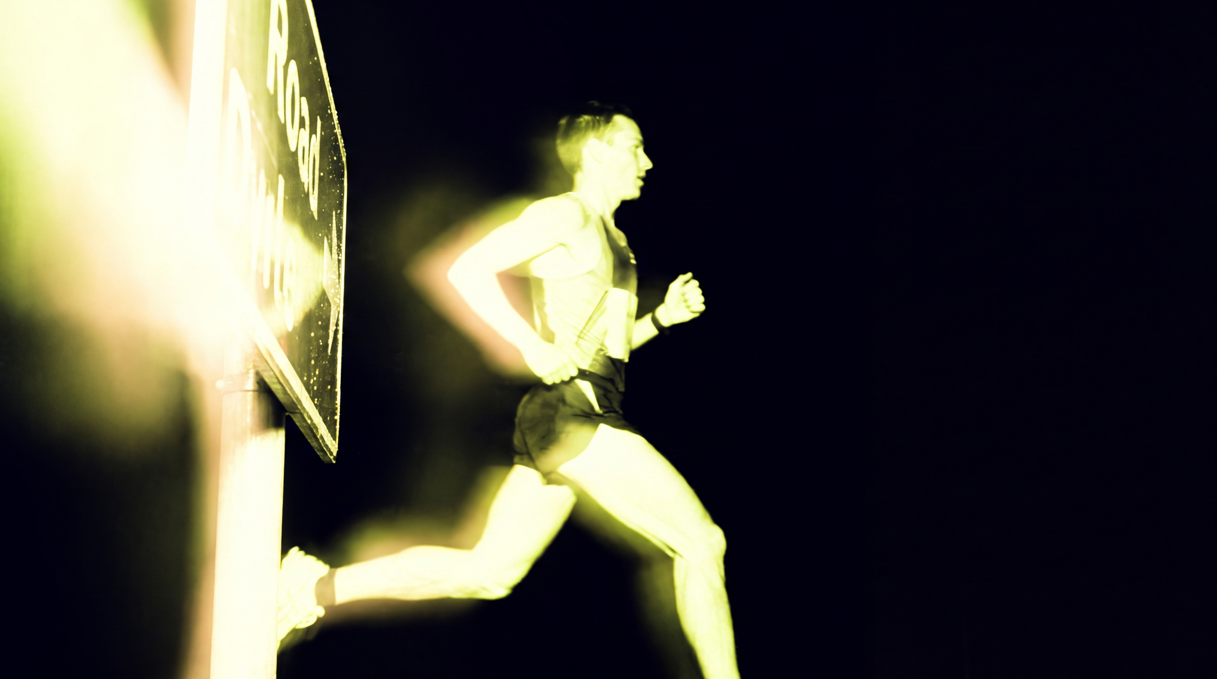 A person running at night, illuminated by a yellowish light, with a black background and partial view of a sign or billboard on the left side.