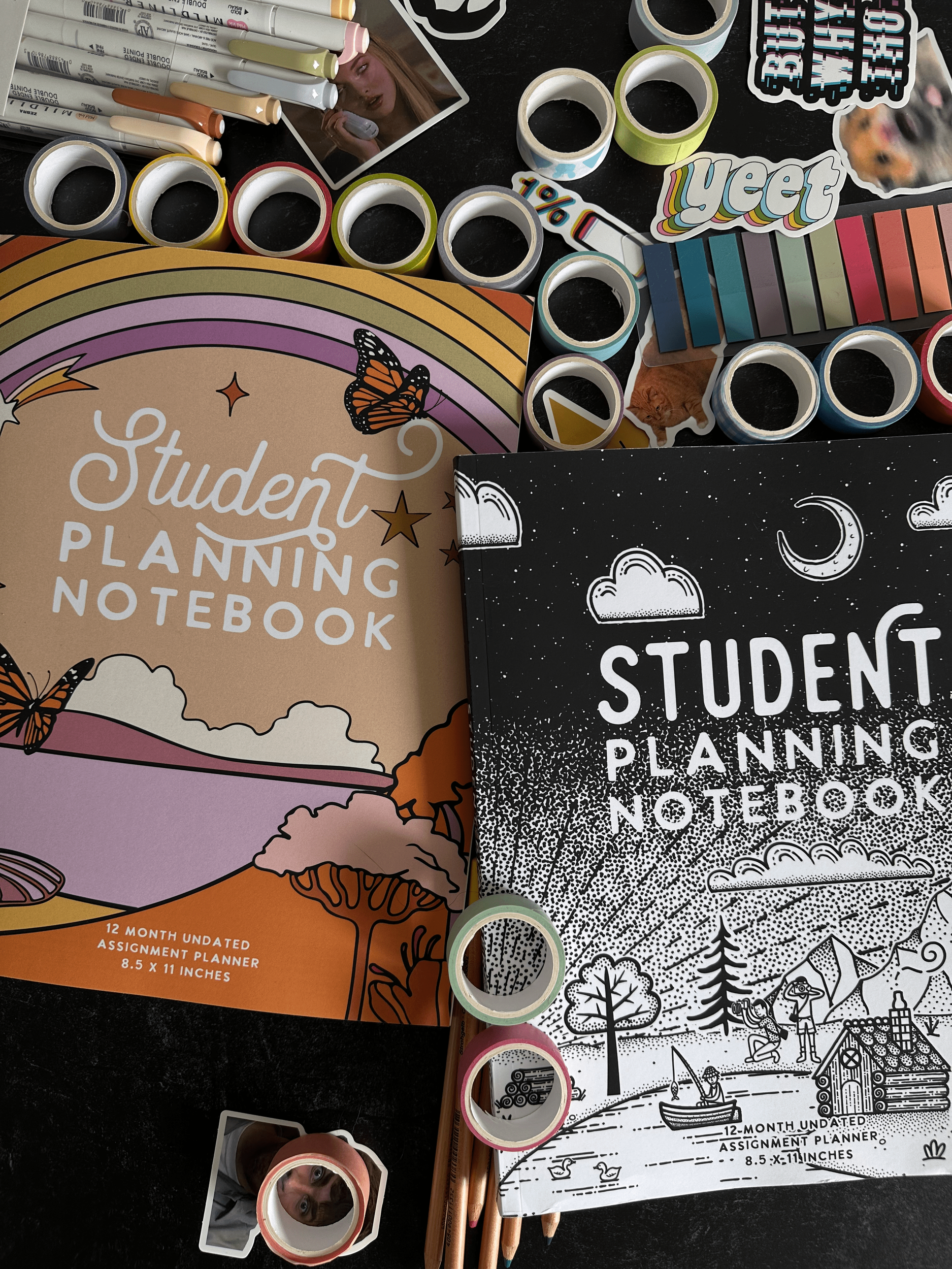 Two student planning notebooks, one in color and one in black and white, surrounded by markers, washi tapes, stickers, and pencils on a black surface.