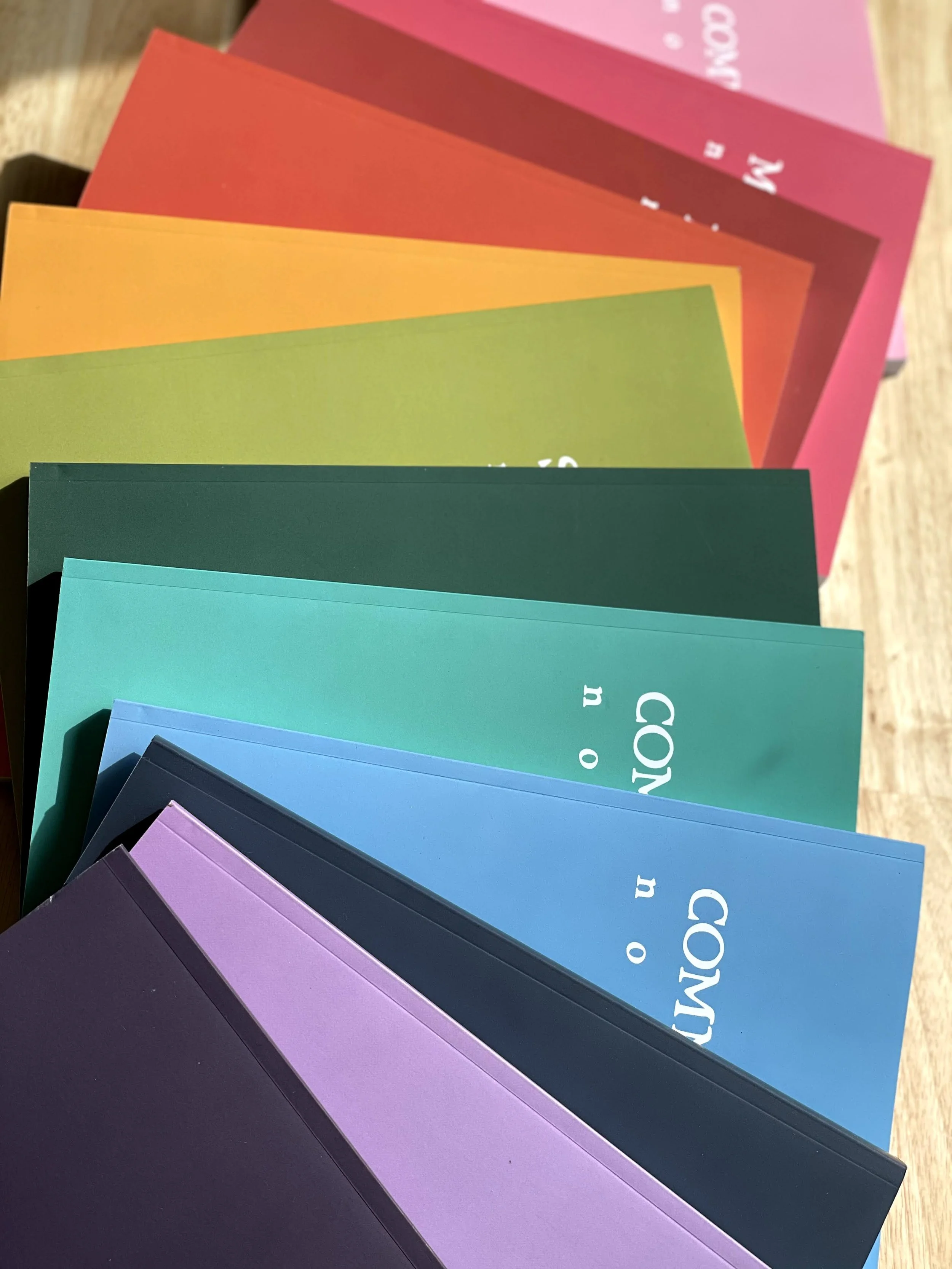 Several colorful envelopes or cards arranged in a fan shape on a wooden surface, in shades of purple, blue, teal, green, yellow, orange, red, and pink.