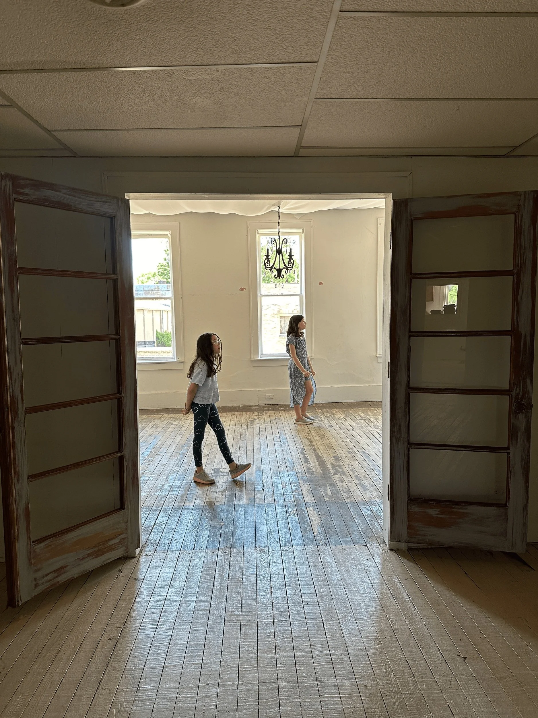 Two young girls walking in a bright room with large windows, a chandelier hanging from the ceiling, and wooden floors, seen through open French doors.