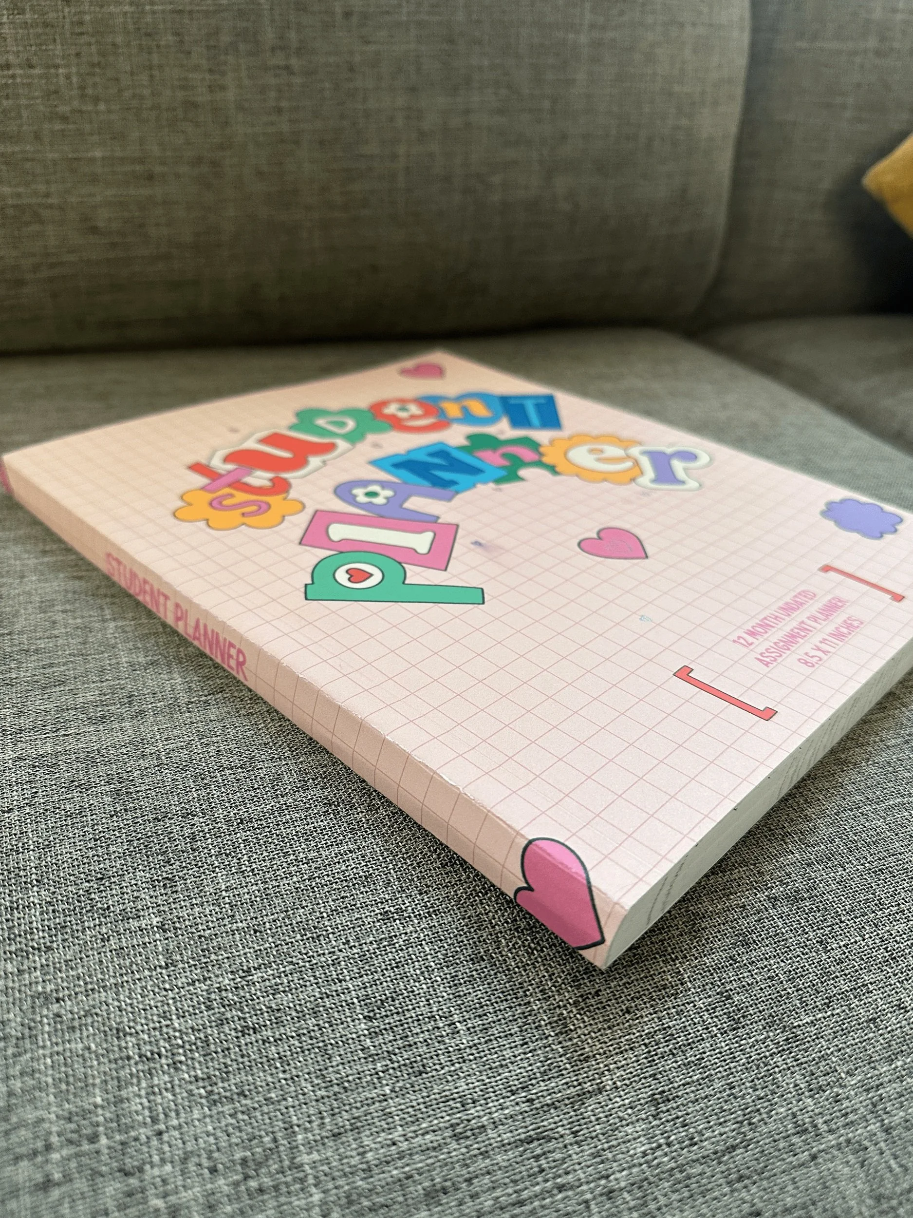 A pink grid-patterned student planner with colorful lettering and hearts on its cover, resting on a grey fabric surface.