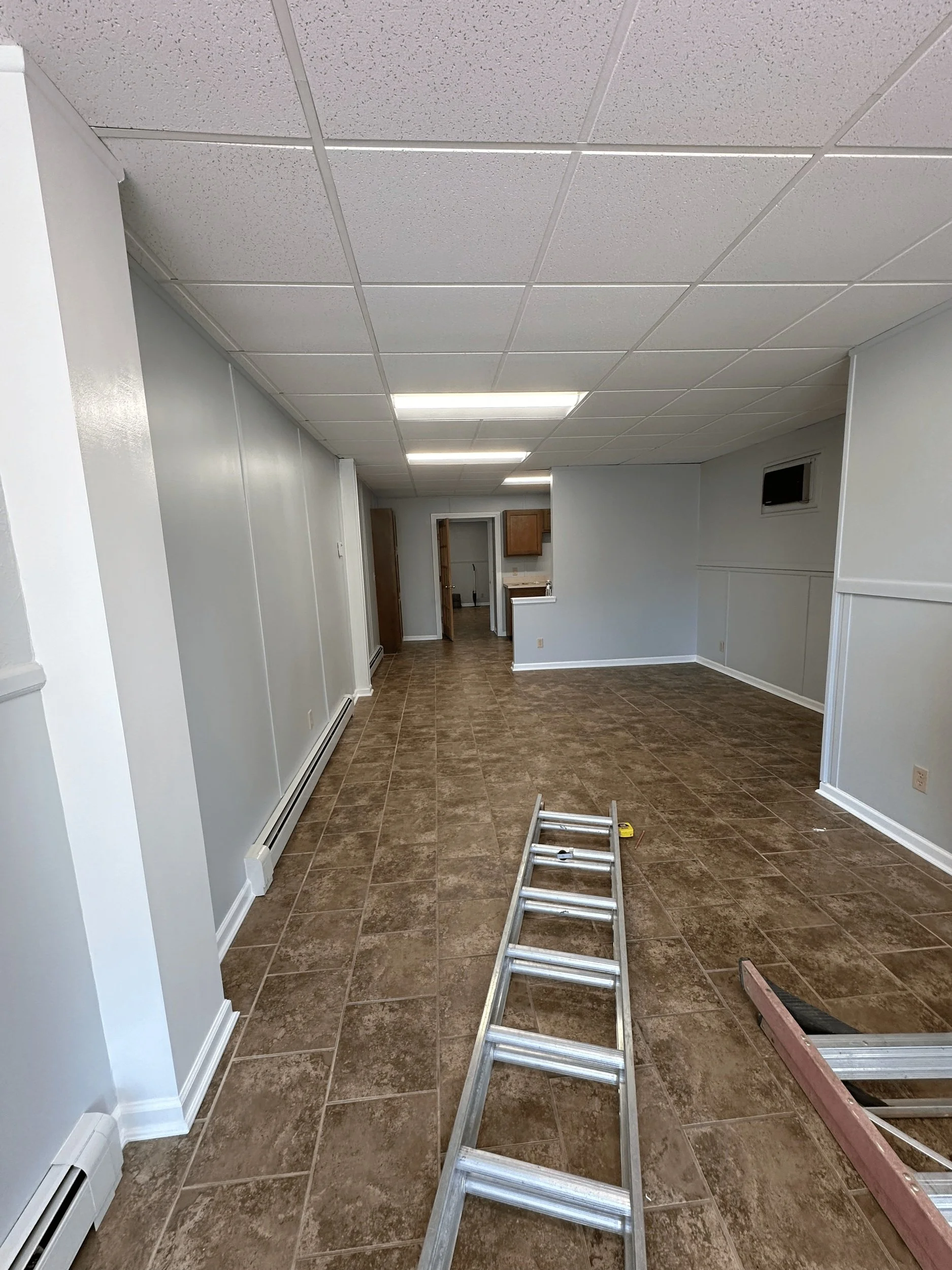 Empty room with tiled floor, white walls, ceiling panels, a ladder, and a small kitchen area in the background with wooden cabinets.