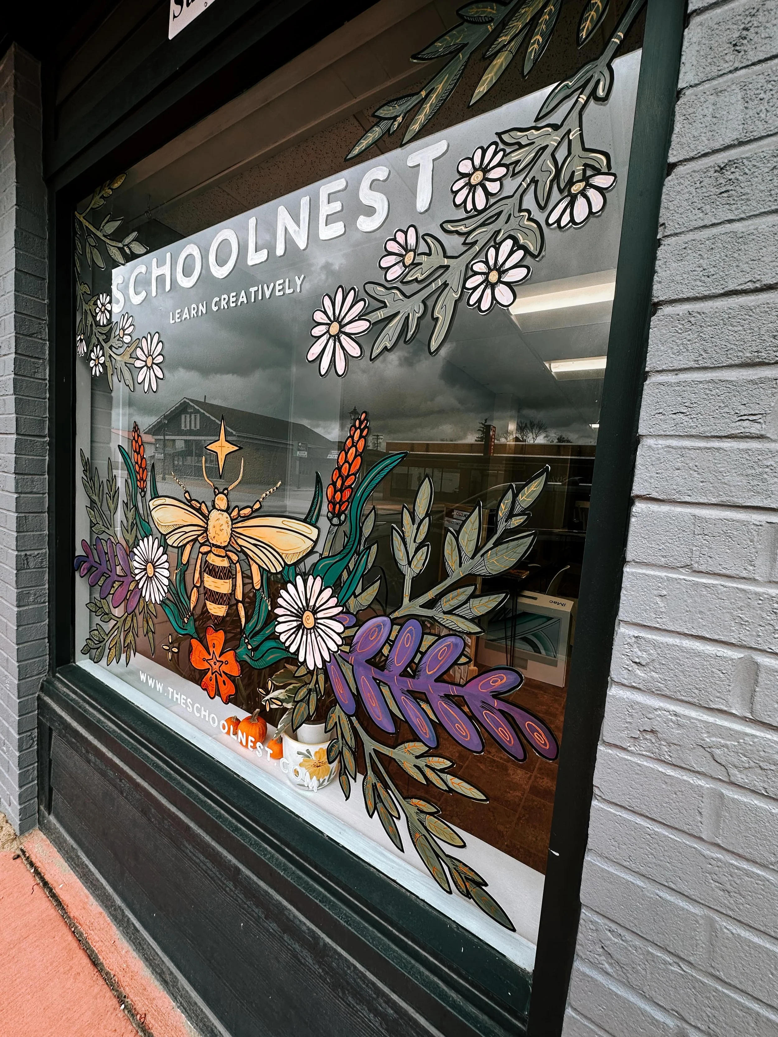 Decorative window display for Schoolnest featuring colorful artwork of a bee, flowers, and foliage with the text 'SCHOOLNEST LEARN CREATIVELY' and the website URL 'WWW.THESCHOOLNEST.COM'.