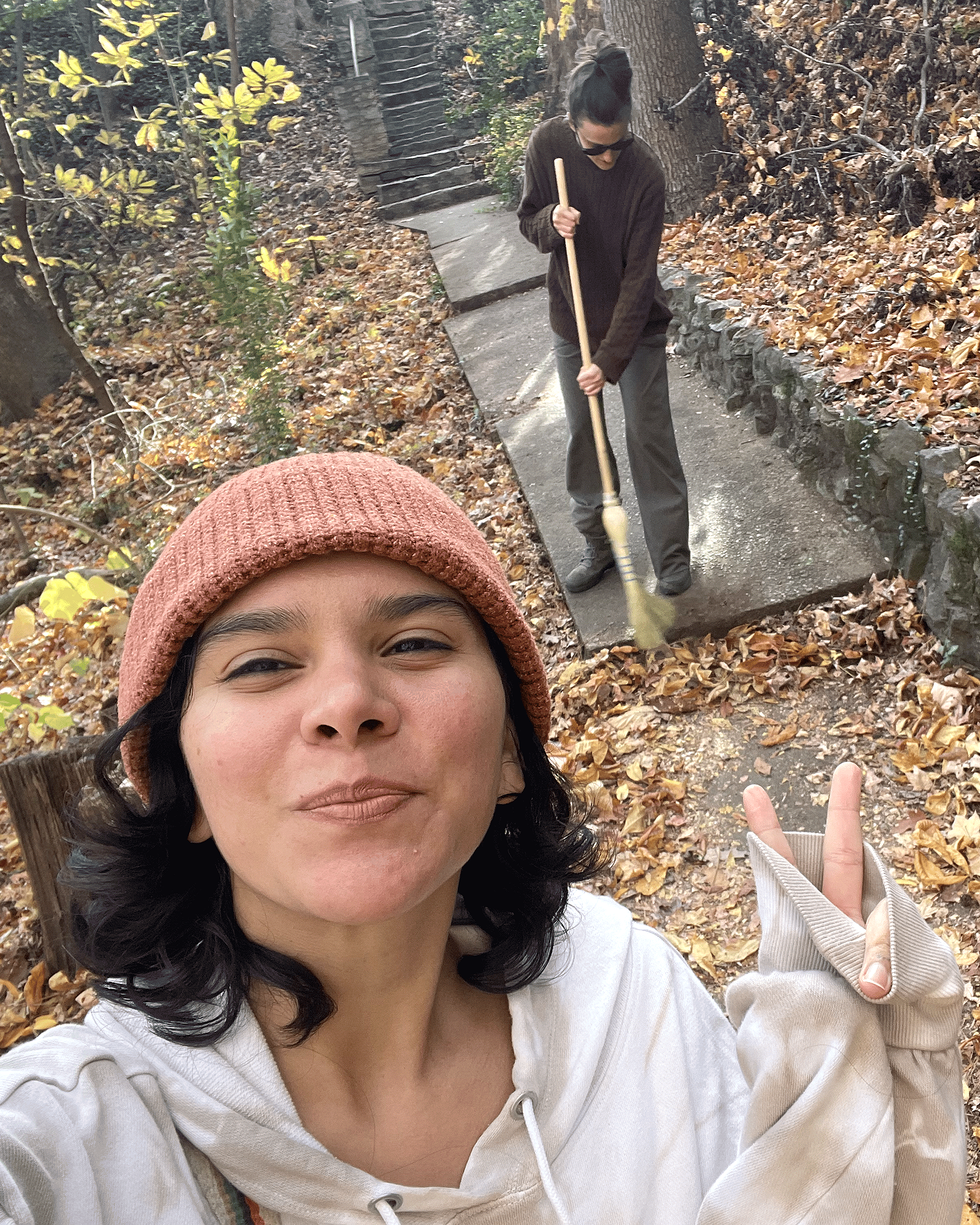 Dalani holds up peace fingers in a beanie as Ala sweeps fallen leaves on a wooded pathway