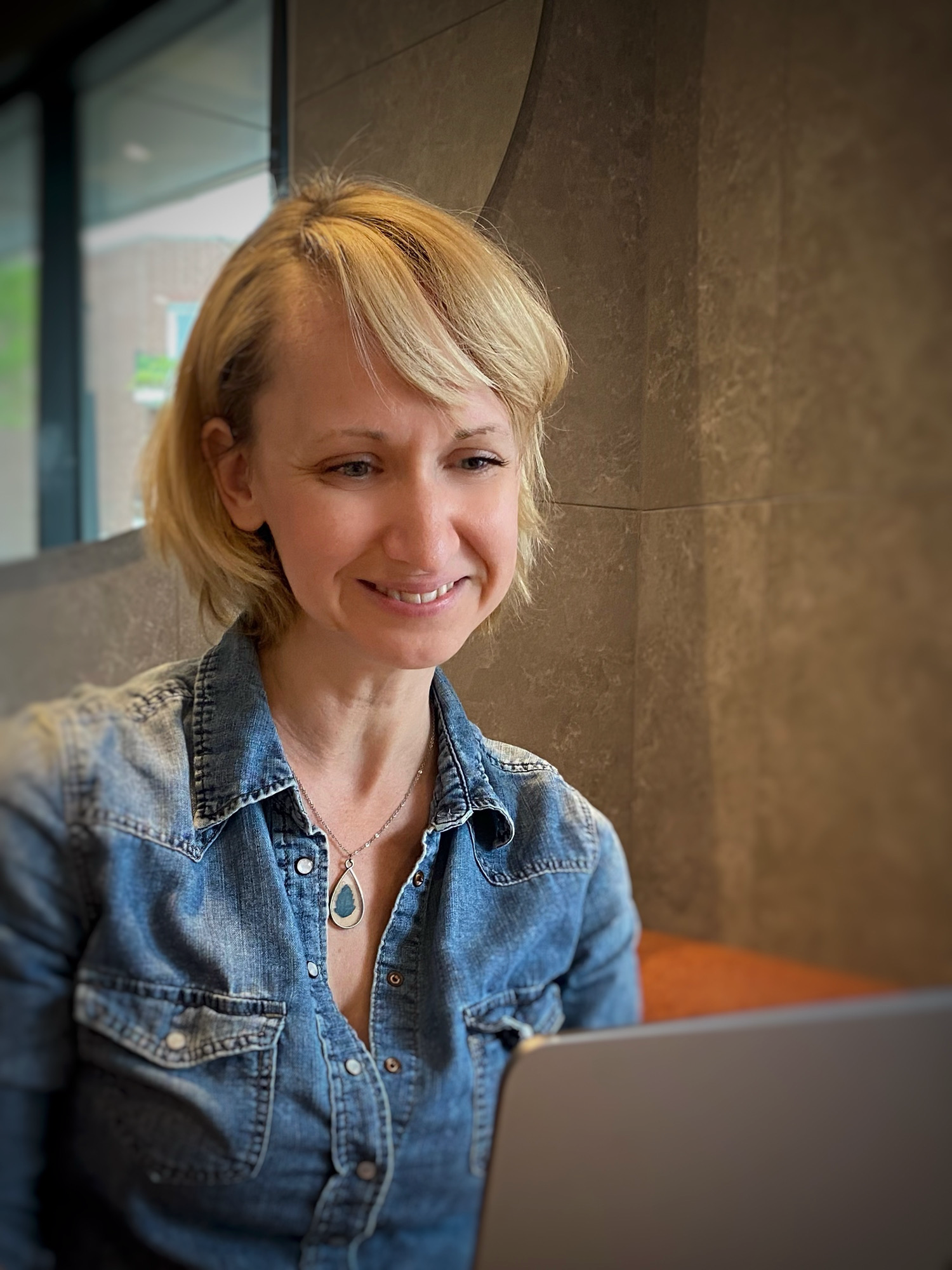 Une femme blonde souriante regarde un ordinateur portable, portant une chemise en jean et un collier avec un pendentif. Elle est assise dans un lieu intérieur avec un mur en pierre et une fenêtre derrière elle.