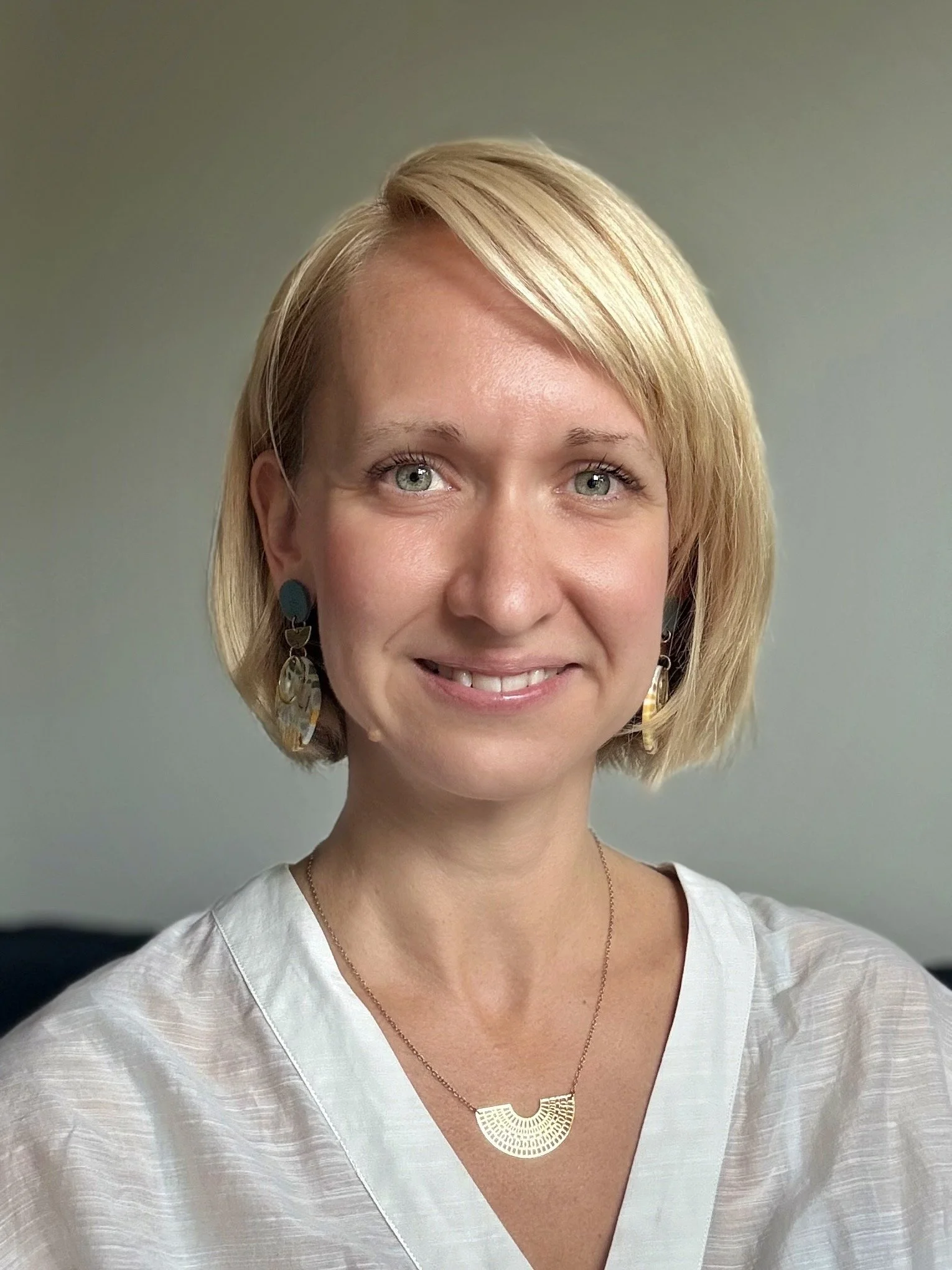 Une femme blonde souriante portant des boucles d'oreilles et un collier, avec un fond neutre.
