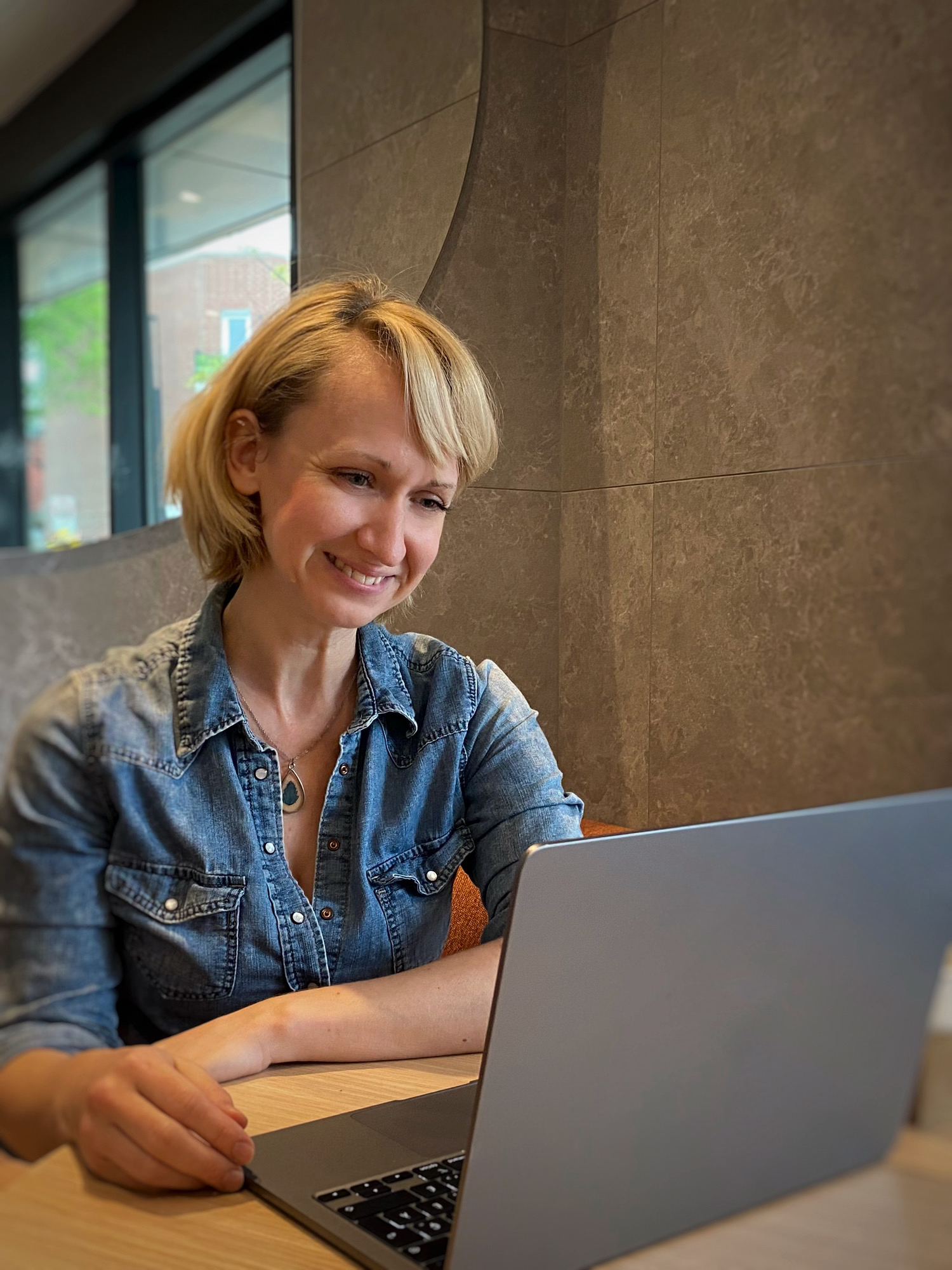 Une femme blonde souriante en veste en jean regarde un ordinateur portable dans un café.