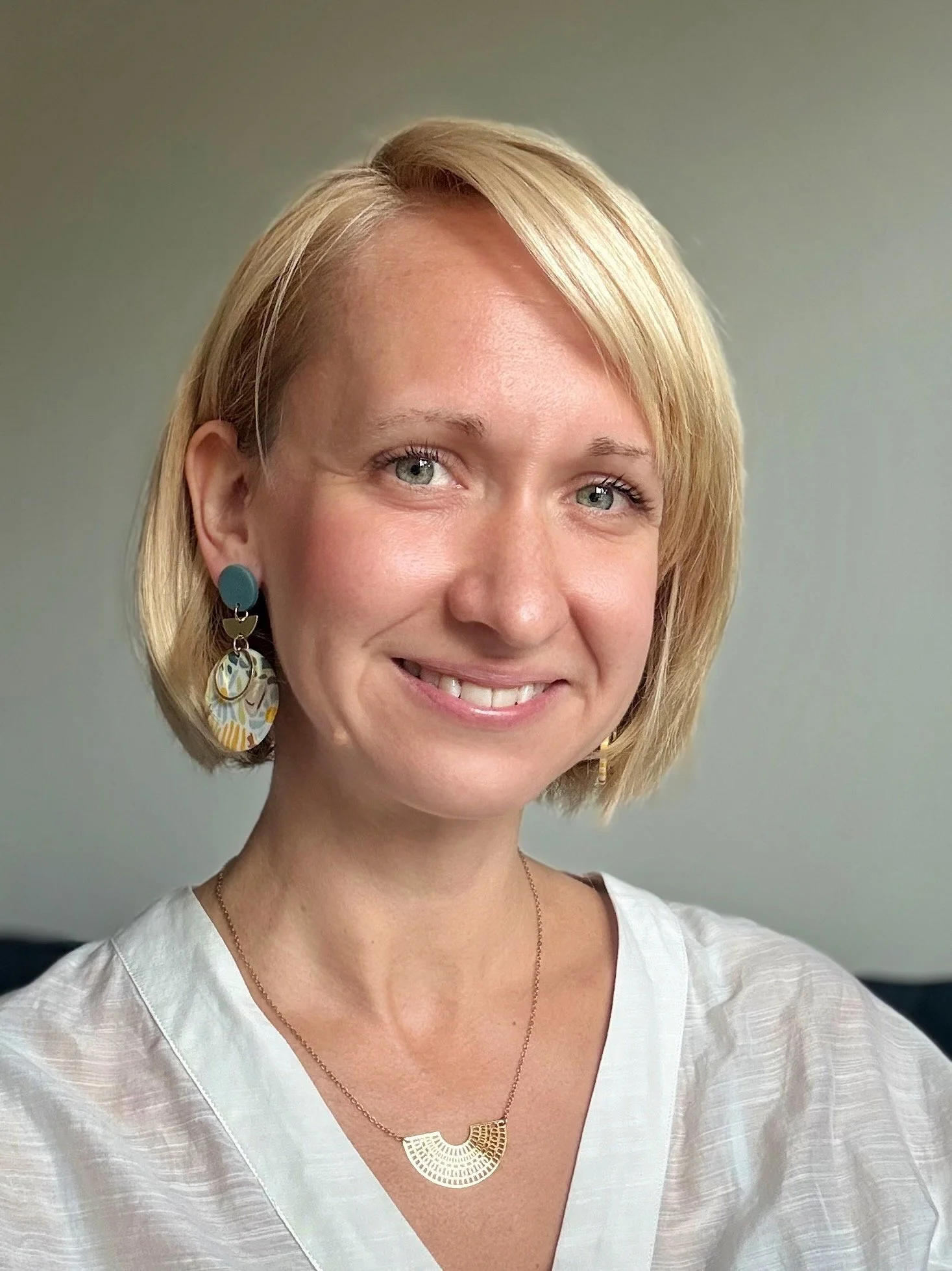 Portrait d'une femme blonde portant des boucles d'oreilles colorées et un collier, souriant devant un fond neutre.