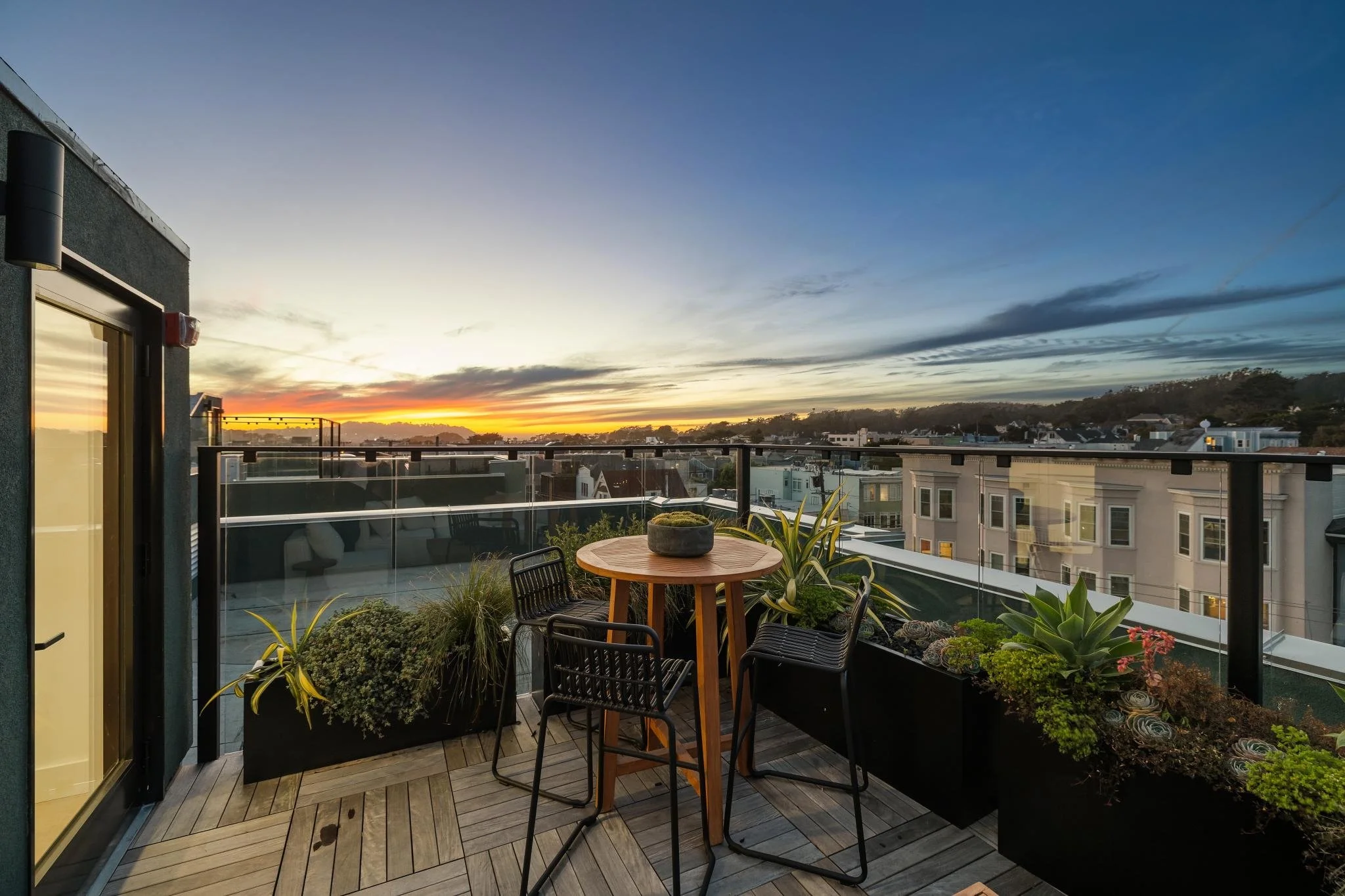 A city rooftop balcony at sunset with a small round wooden table, three black chairs, and rectangular planters filled with various succulents and plants, overlooking neighboring buildings and a colorful sky with clouds.