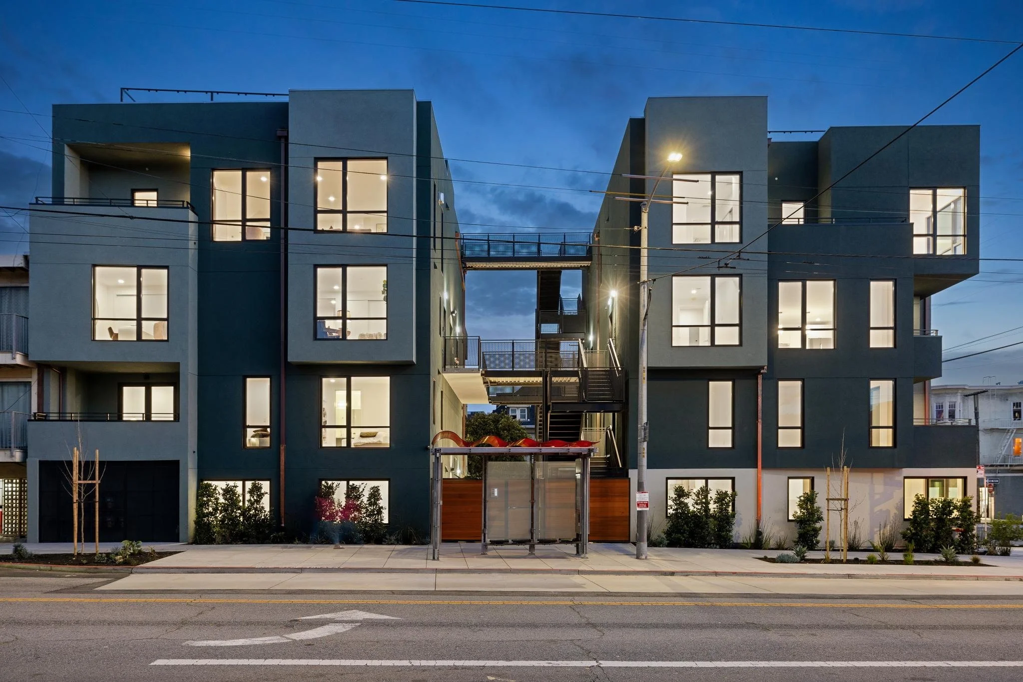 Modern multi-story apartment building with illuminated windows, exterior staircases, and landscaped surroundings at dusk.
