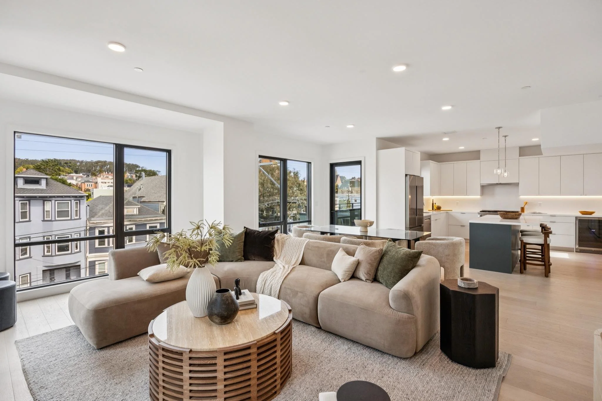 Modern open-concept living room and kitchen with large windows, beige sectional sofa with pillows, coffee table with decorative vases, and white minimalist kitchen with island, stainless steel refrigerator, and pendant lighting.