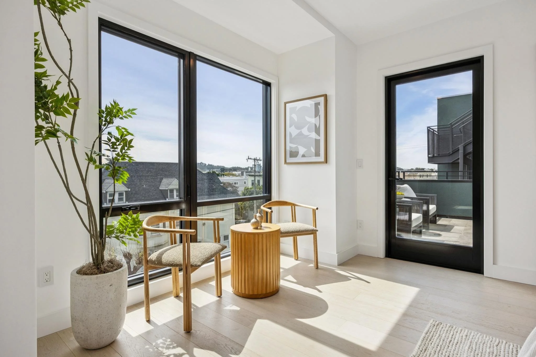 Modern interior with a potted plant, two wooden chairs, a small wooden side table with decorative objects, large windows, and a balcony door, with sunlight streaming in.