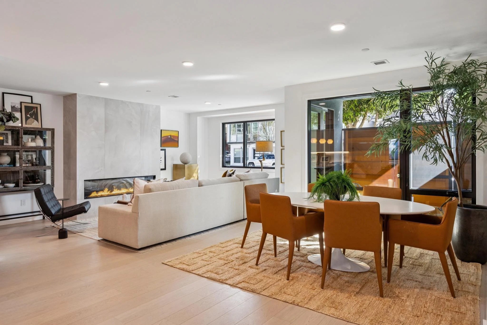 Modern living room with white walls, light wood flooring, a white sofa, a round dining table with six orange chairs, large windows, potted plants, and a built-in fireplace.