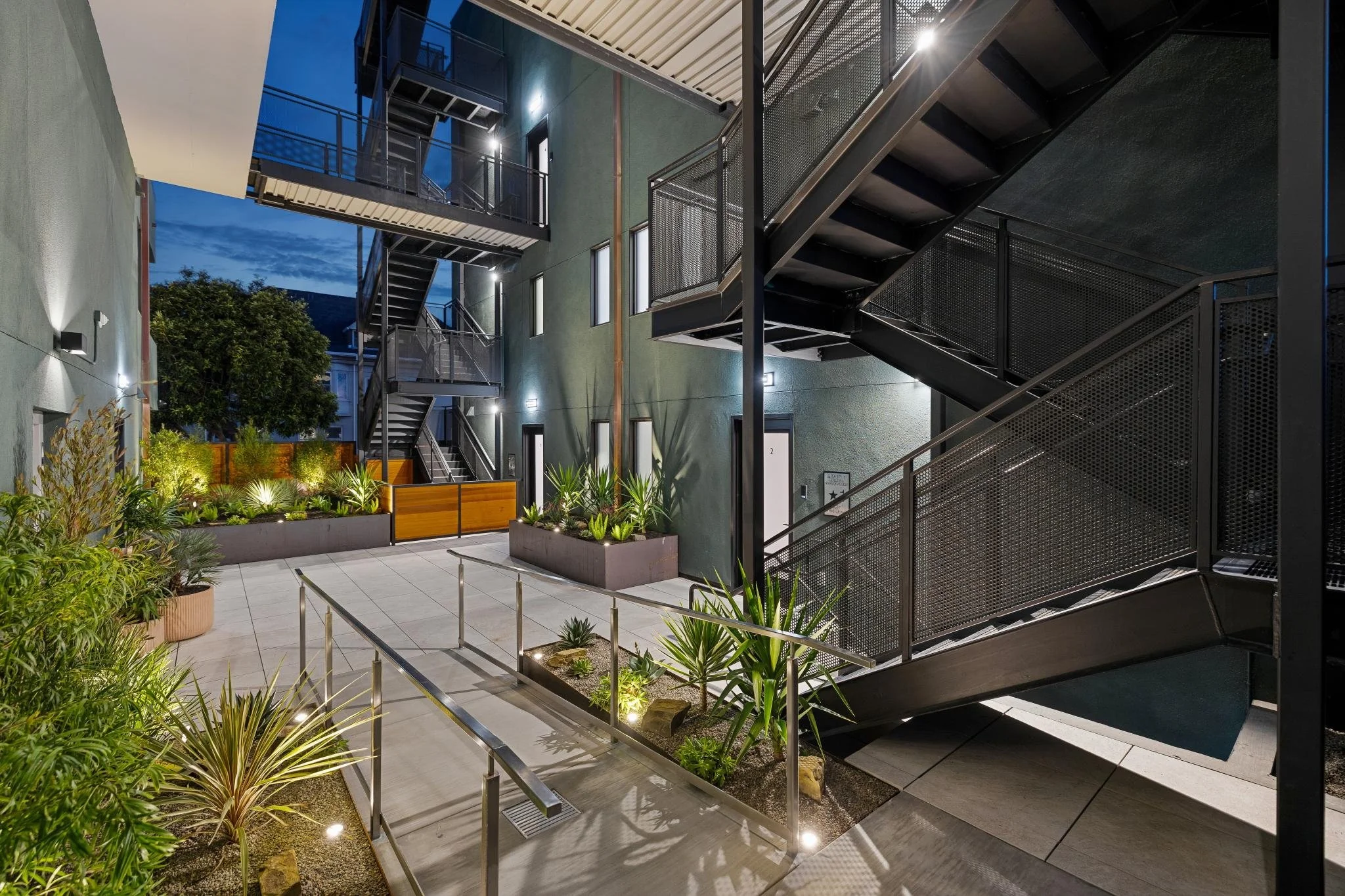 Modern apartment complex courtyard with metal stairs, potted plants, outdoor lighting, and landscaped greenery at dusk.