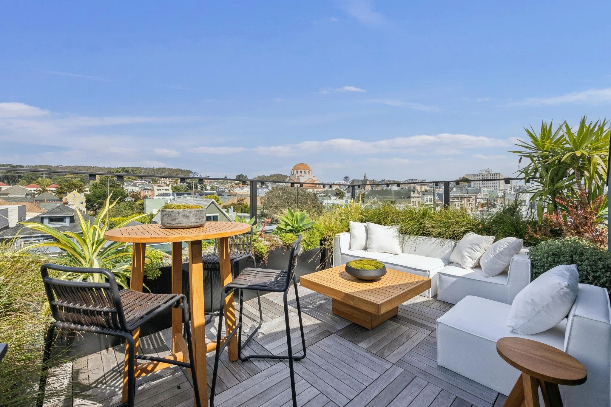 Urban rooftop patio with outdoor white sofa, wooden tables, black chairs, and potted plants overlooking cityscape with blue sky.