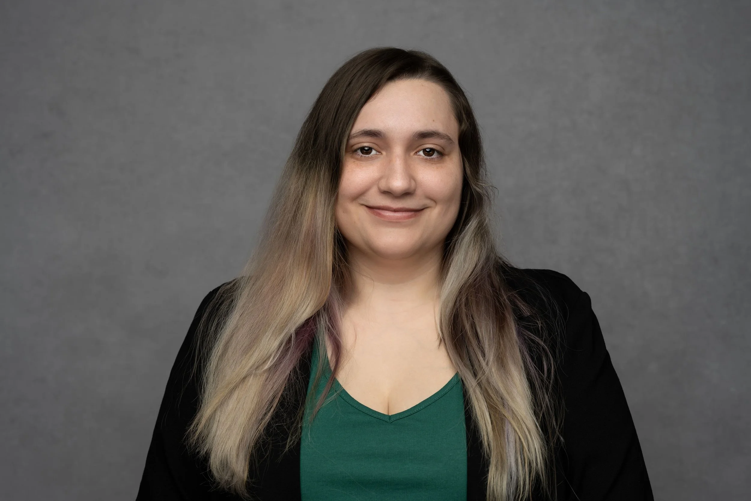 A woman with long, lightly colored hair, wearing a black blazer over a green top, smiling against a gray background.