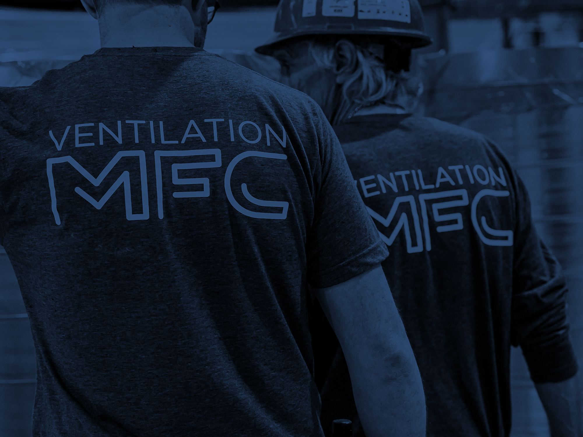 Deux personnes portant des t-shirts gris avec le texte "VENTILATION MEC" et portant des casques de chantier, vues de dos, dans un environnement industriel ou de construction.