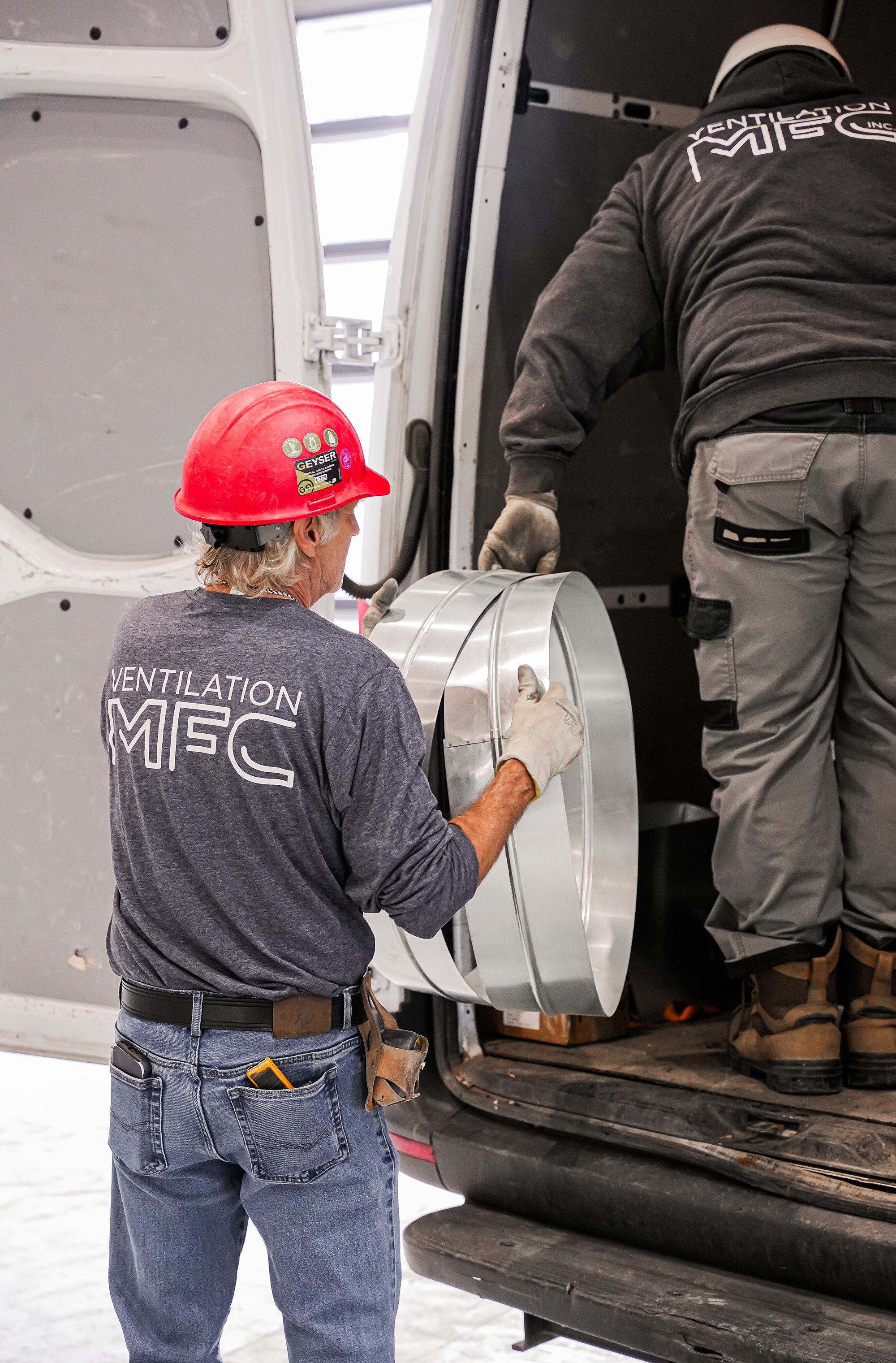 Deux hommes travaillent ensemble dans une cabine de véhicule, l'un porte un casque rouge et un t-shirt gris avec le logo 'VENTILATION MFC', l'autre homme, vêtu de gris, est à l'intérieur du véhicule, semblant installer ou réparer quelque chose.