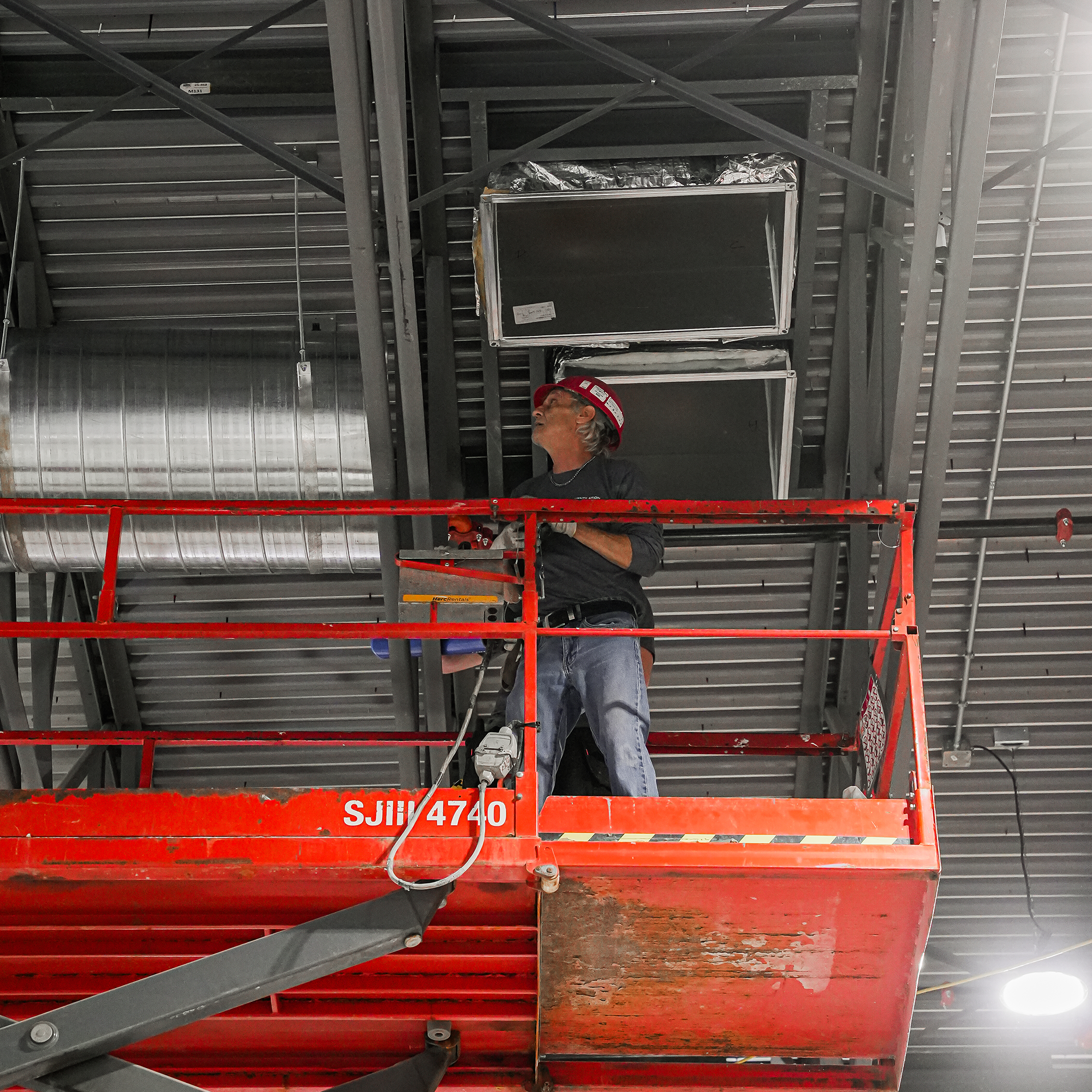 Un homme portant un casque de sécurité rouge et des gants travaille à l'intérieur d'une structure métallique, utilisant une échelle pour atteindre une partie supérieure de l'installation.