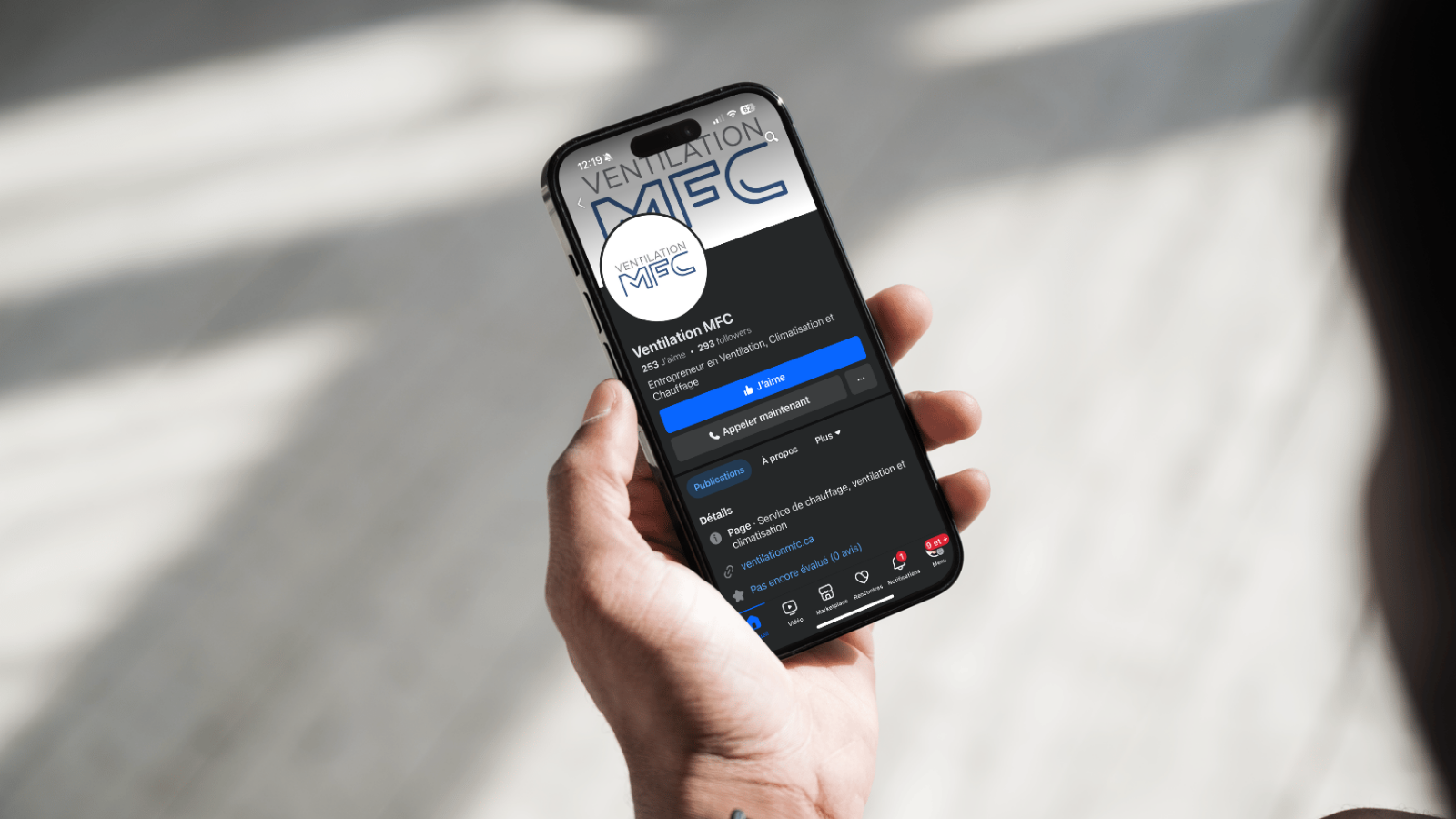 Une personne tenant un smartphone affichant la page Facebook d'une entreprise de ventilation, chauffage et climatisation nommée Ventilation MFC.