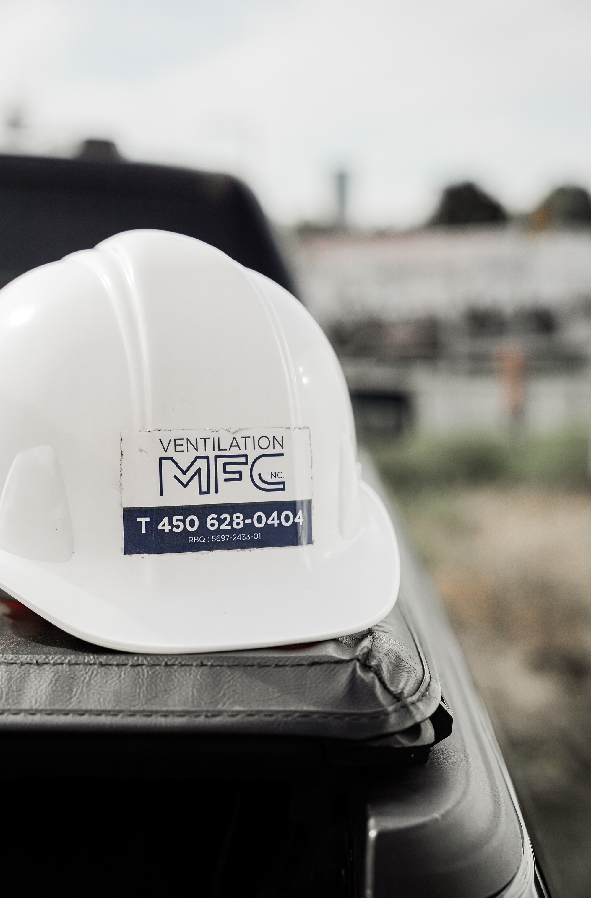 Casque de sécurité blanc avec une étiquette indiquant ventilation MFC, posé sur une surface en tissu, avec un véhicule en arrière-plan