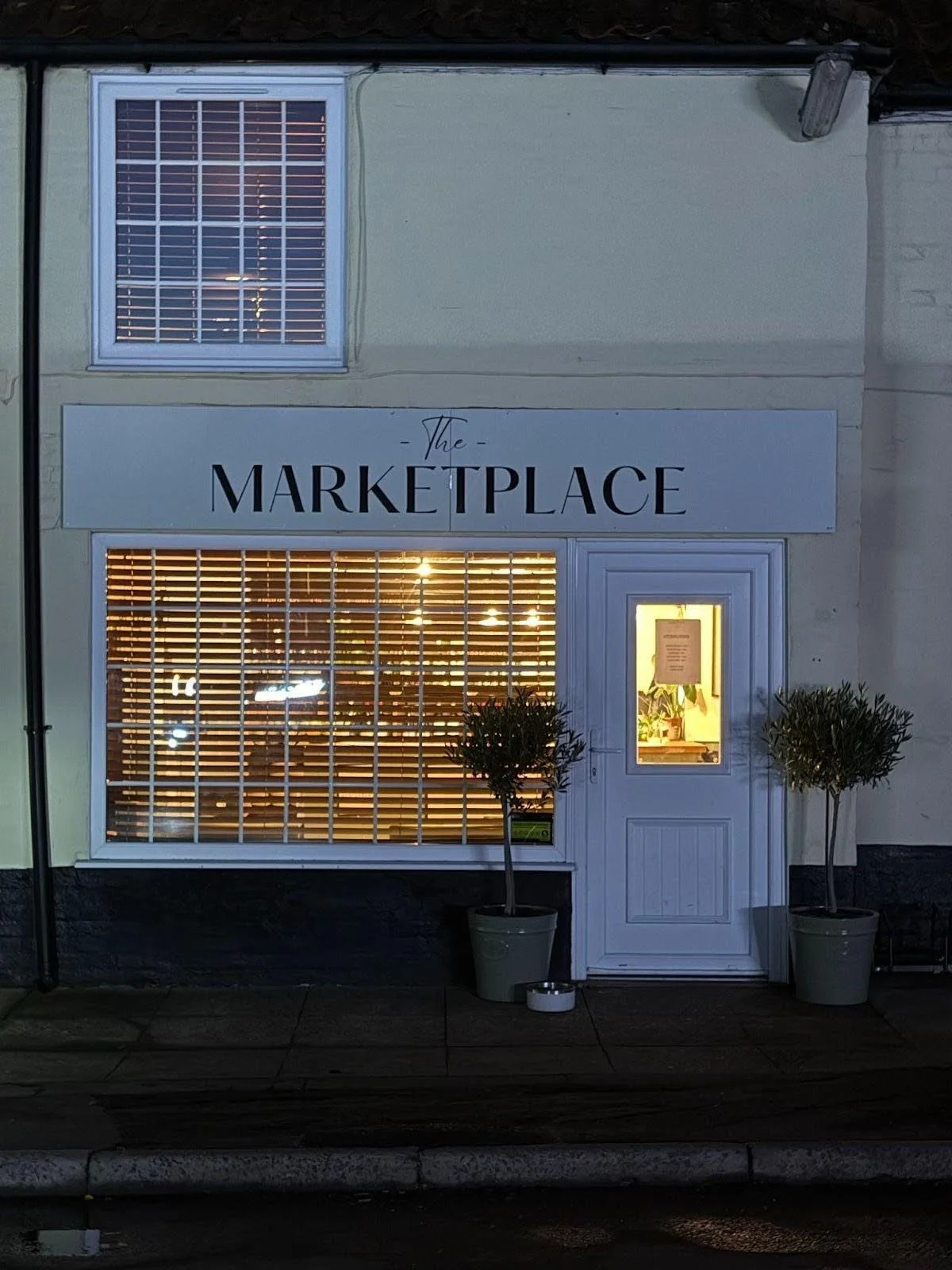 Two people in winter clothing on ladders fixing cardboard over a storefront with a neon sign. A white van and recycling bin are visible.