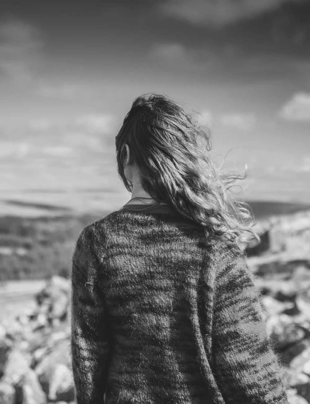 A woman with wavy hair stands outdoors, facing away from the camera, with cloudy skies and a landscape in the background.