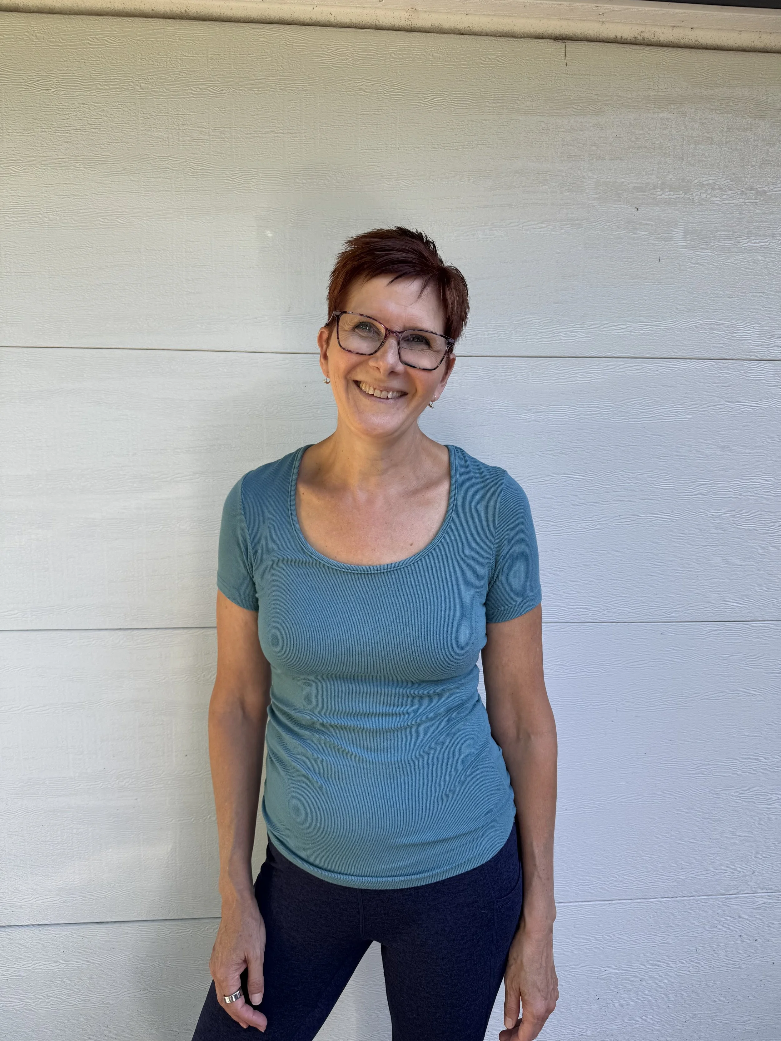 A smiling woman with short, reddish-brown hair and glasses standing in front of a beige wall with horizontal lines, wearing a teal short-sleeved top and dark pants.