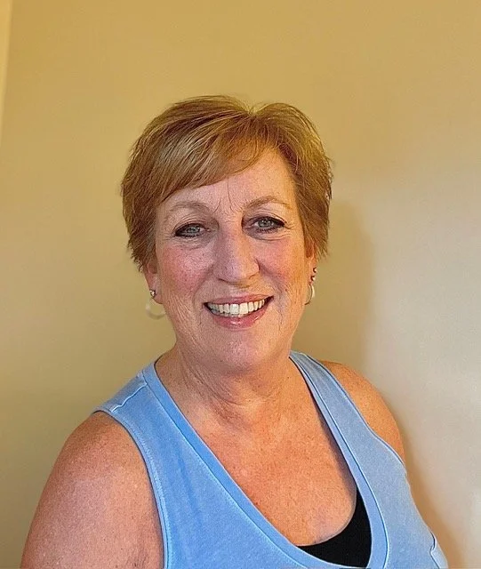 A smiling woman with short, light brown hair, wearing a blue sleeveless top, standing against a plain yellow wall.