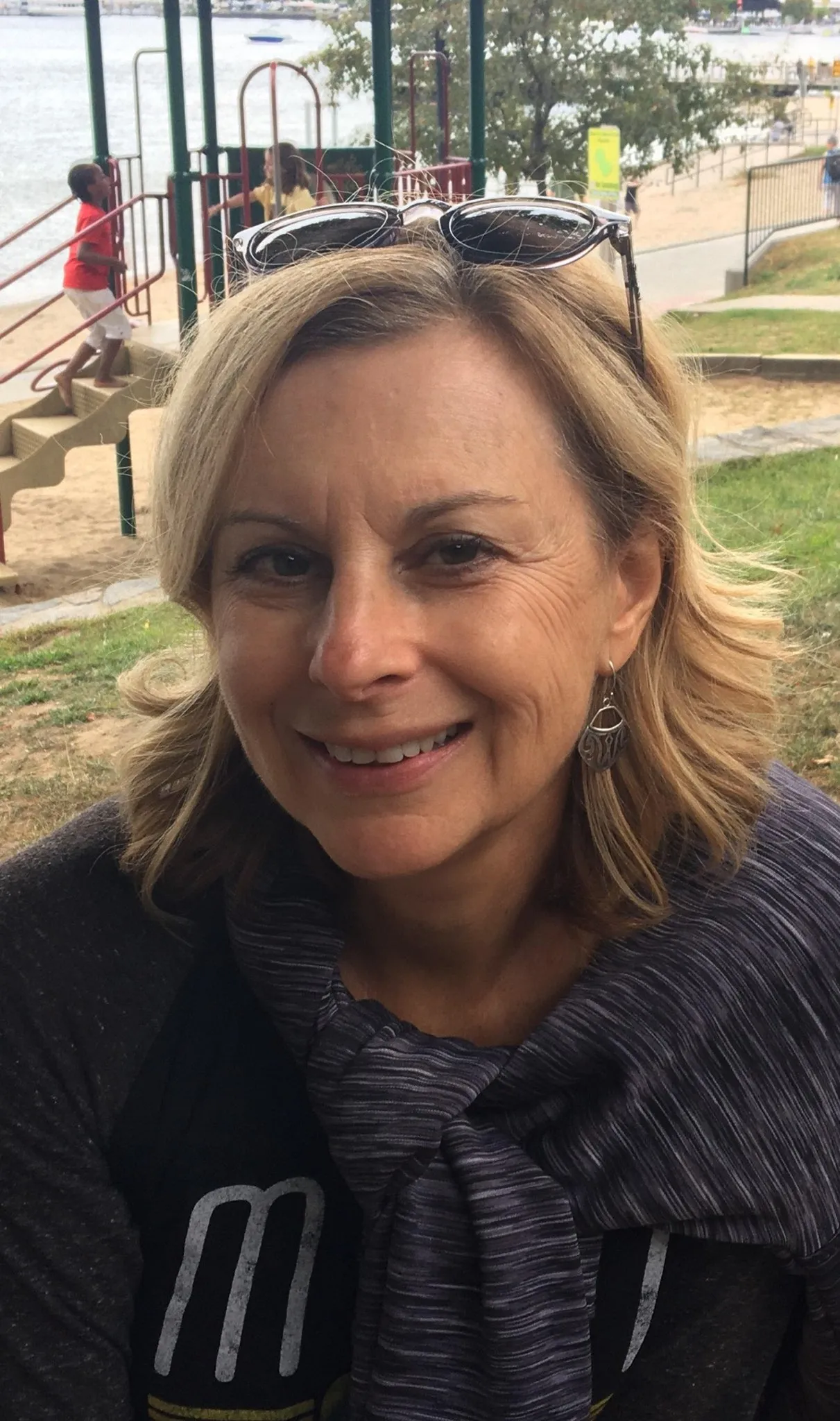 Close-up of a smiling woman with sunglasses on her head, outdoors near a park with playground equipment and a body of water in the background.