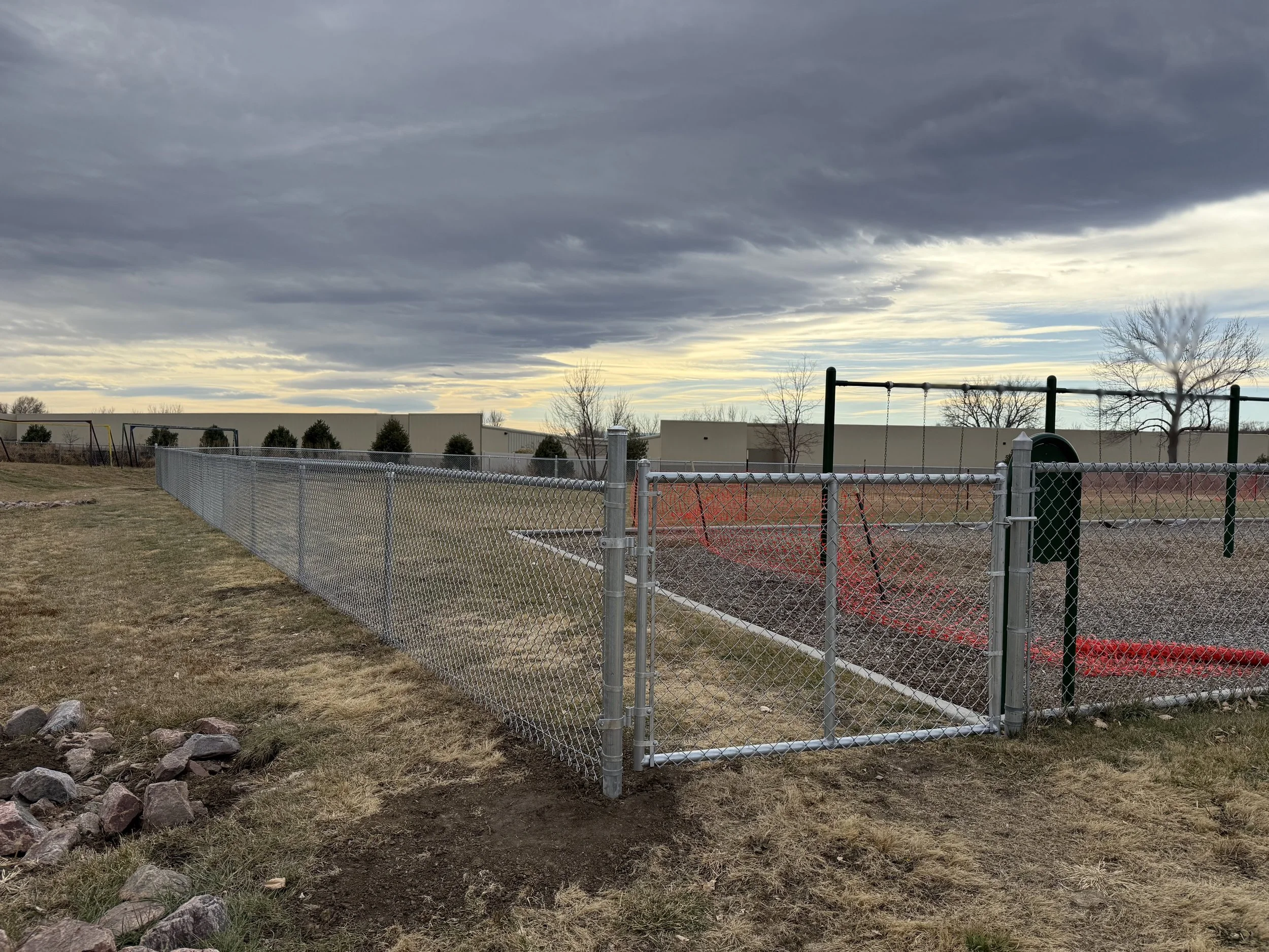 Empty fenced sports field with a swing set and a large building in the background under a cloudy sky.