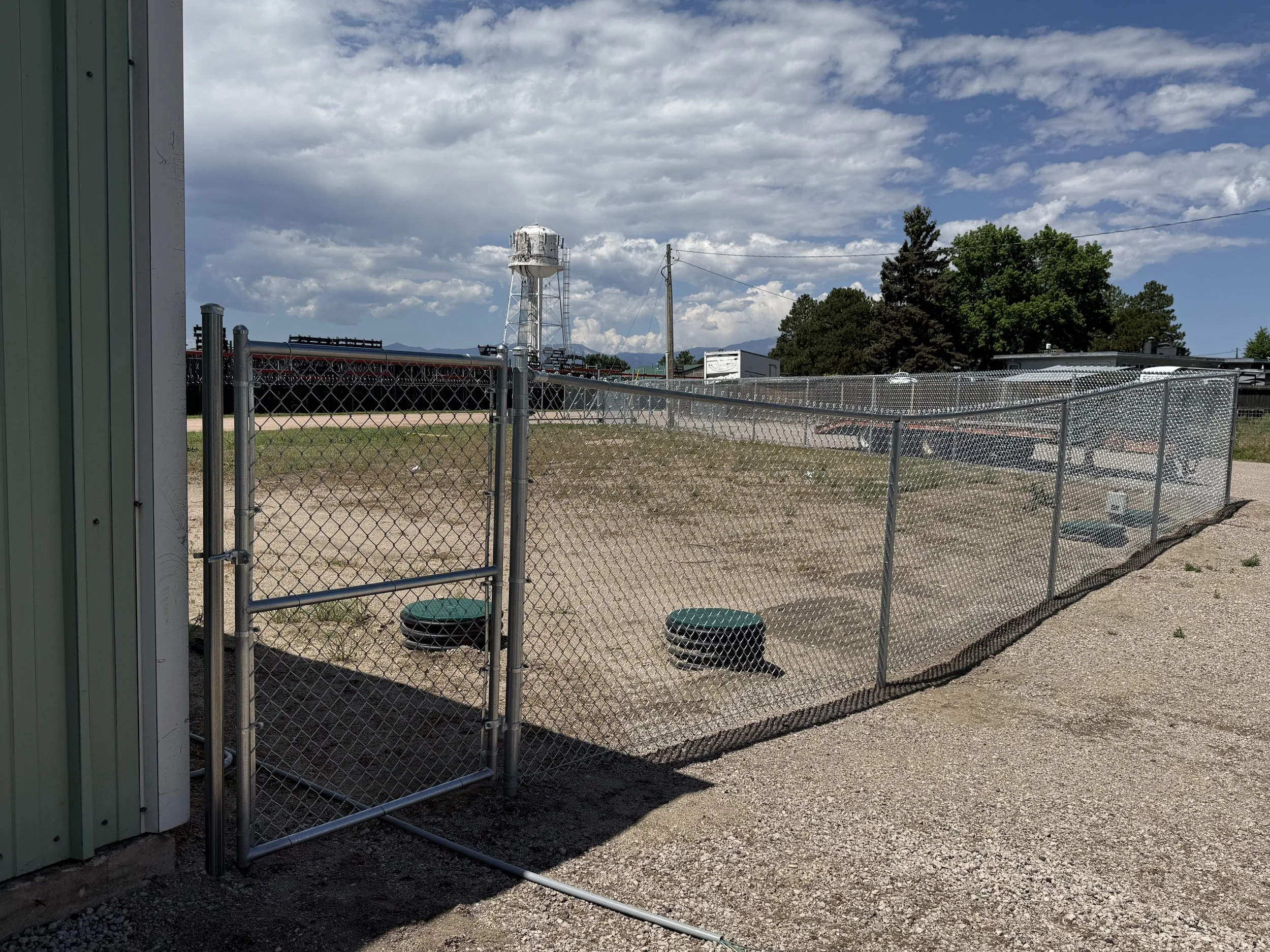 A fenced-off area with a small door, two green water bowls, and a dirt ground, with trees, industrial buildings, and a water tower in the background under a partly cloudy sky.