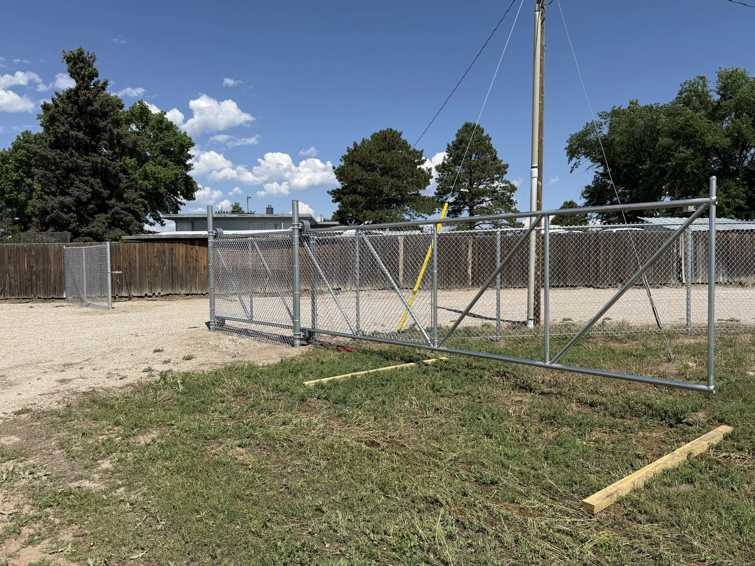 A detached metal gate leaning against a utility pole in a yard, with a wooden fence, trees, and a house in the background on a sunny day.