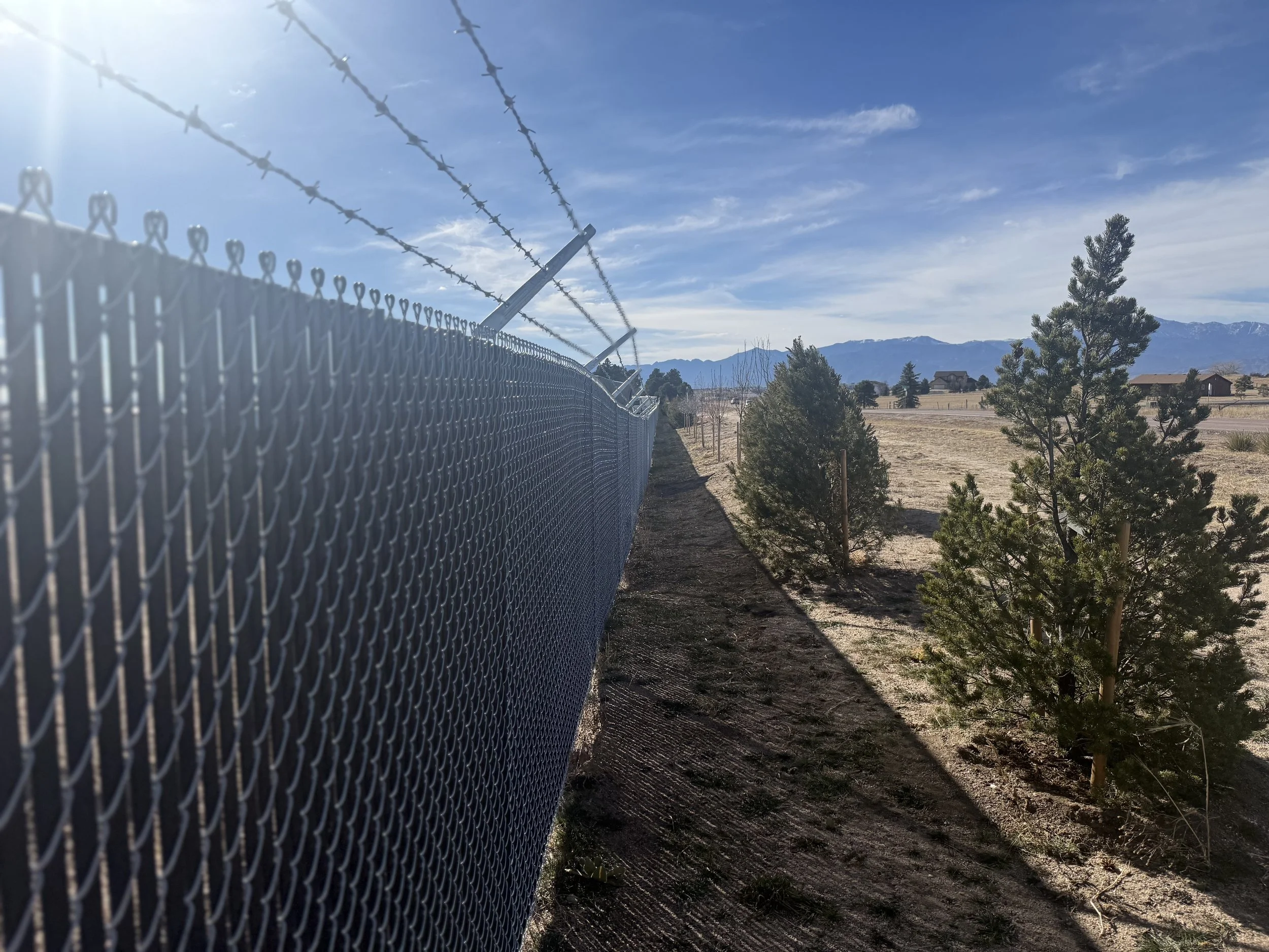 A chain-link fence topped with barbed wire running alongside small trees and open land, with mountains in the distance under a blue sky.