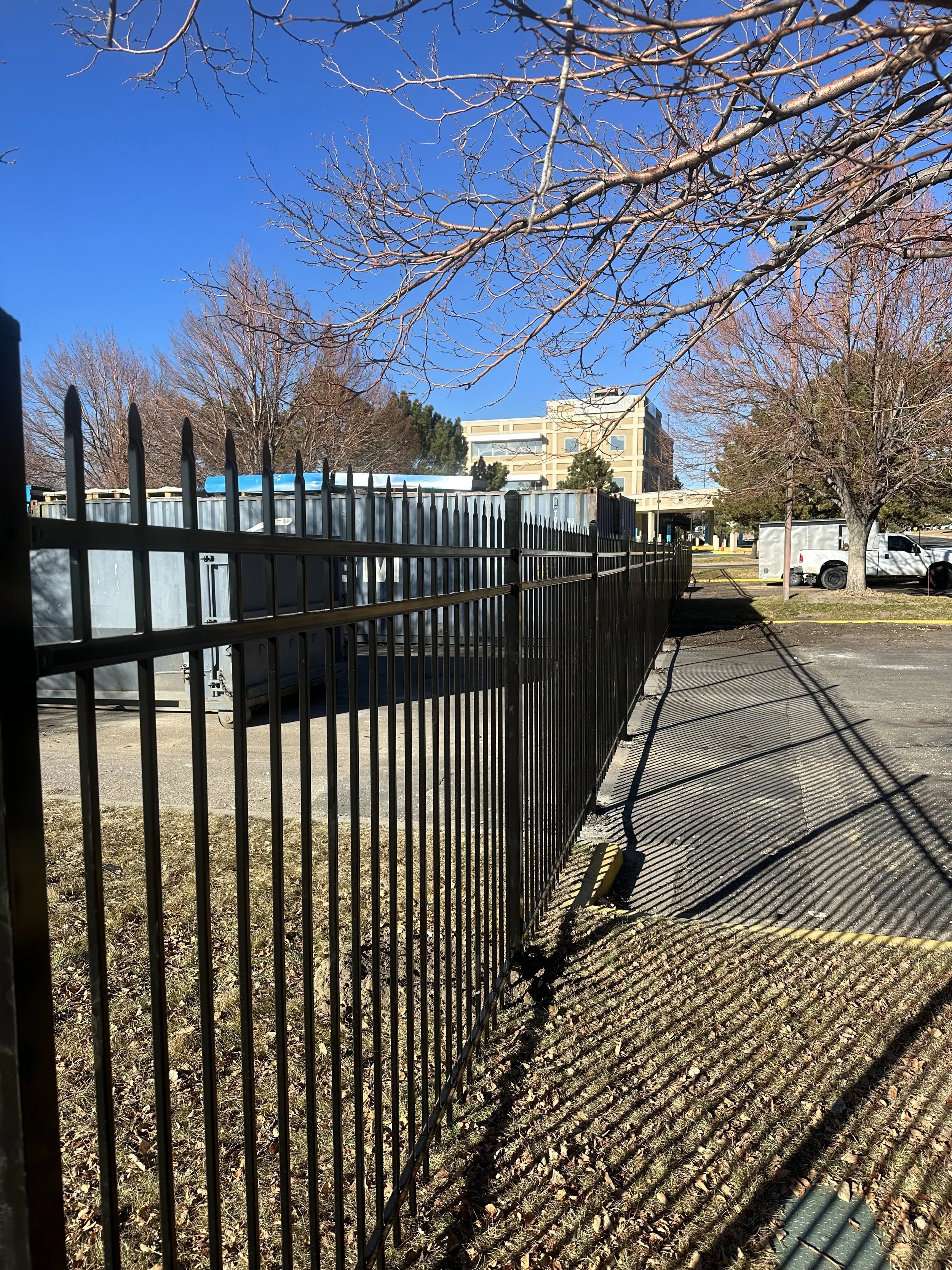 A black metal fence casting striped shadows on the sidewalk next to a parking lot with a few parked vehicles and leafless trees under a clear blue sky.