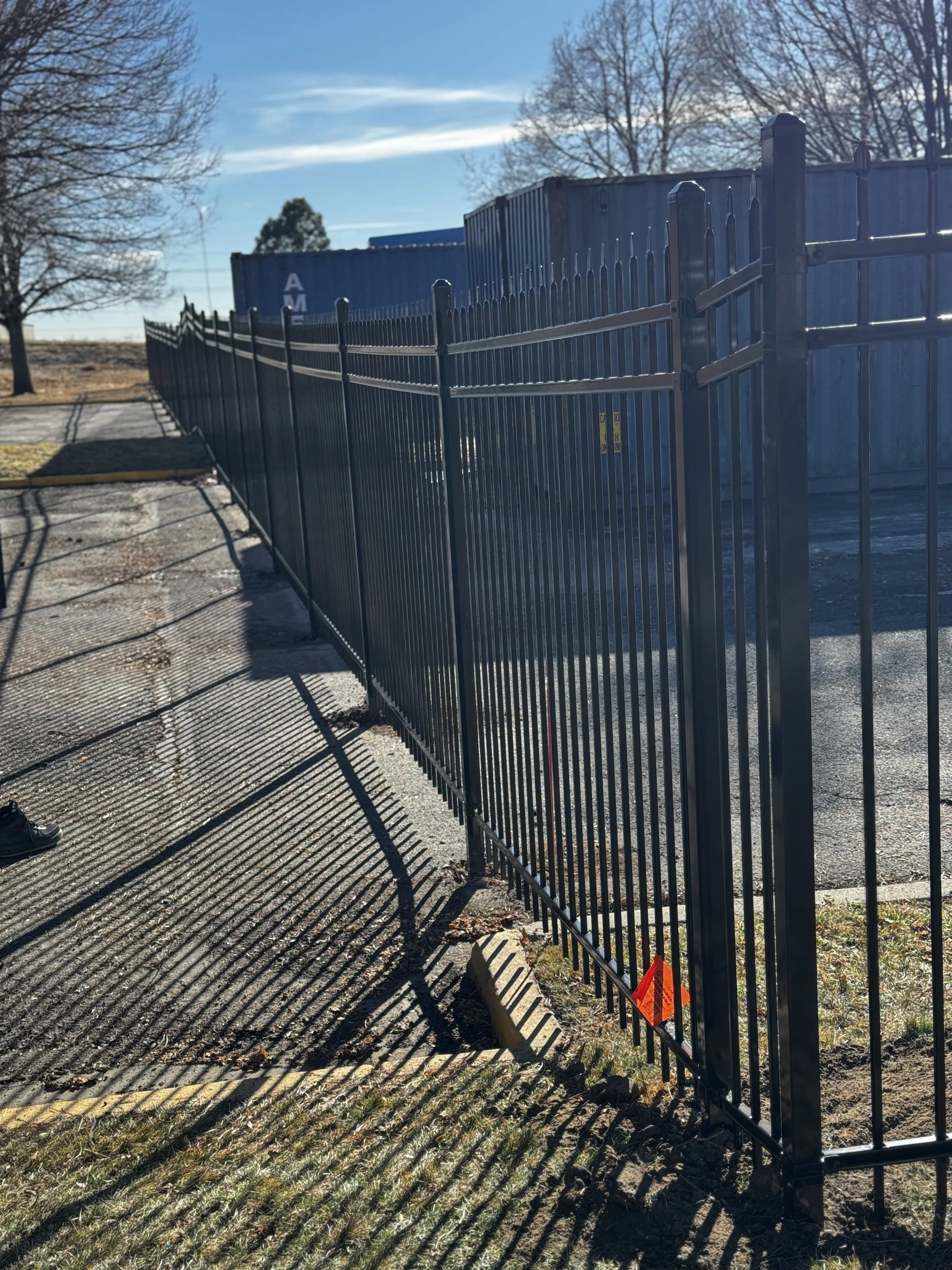 Black metal fence casting shadows on sidewalk next to parking lot with containers and trees in the background, clear blue sky.