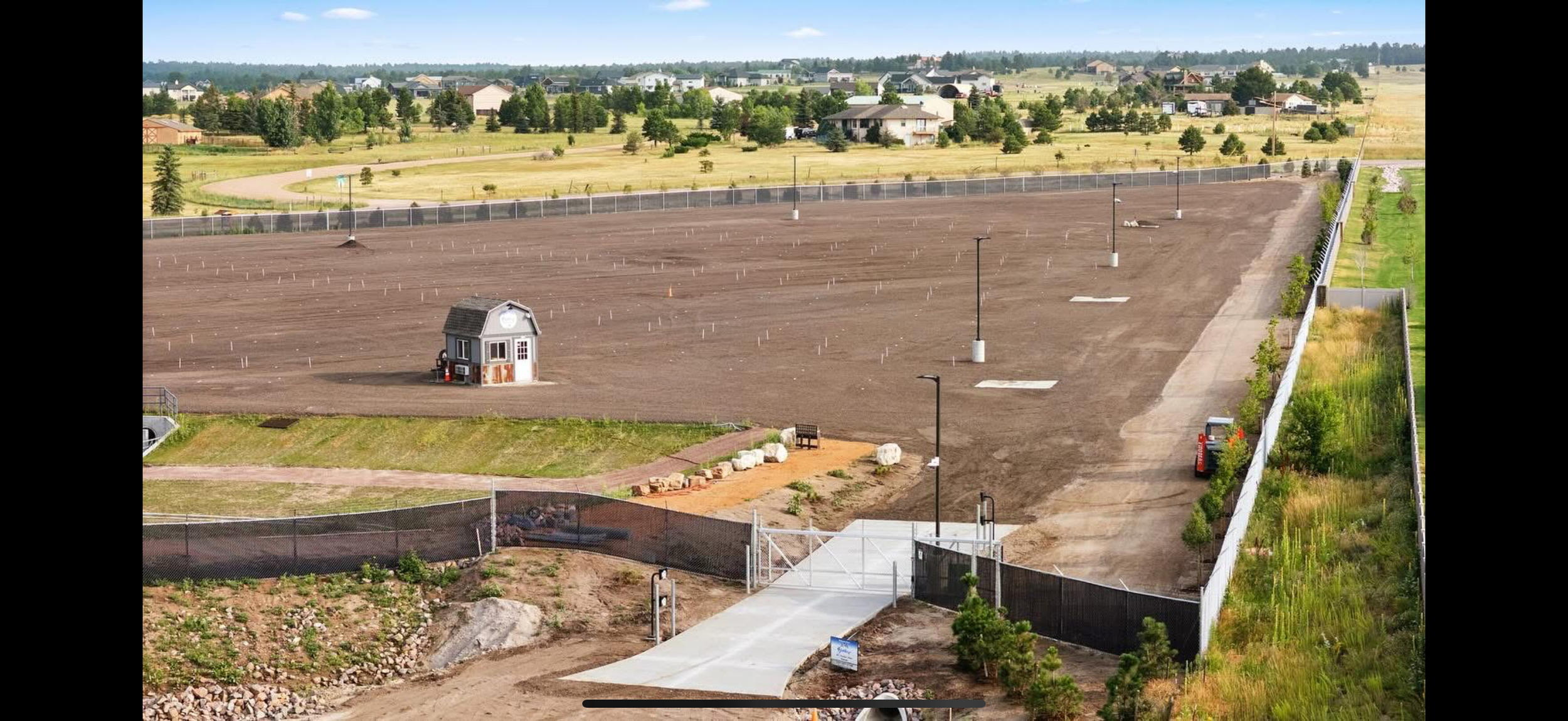 A fenced yard with a small house, surrounding trees, and a large, cleared dirt lot with evenly spaced light poles.