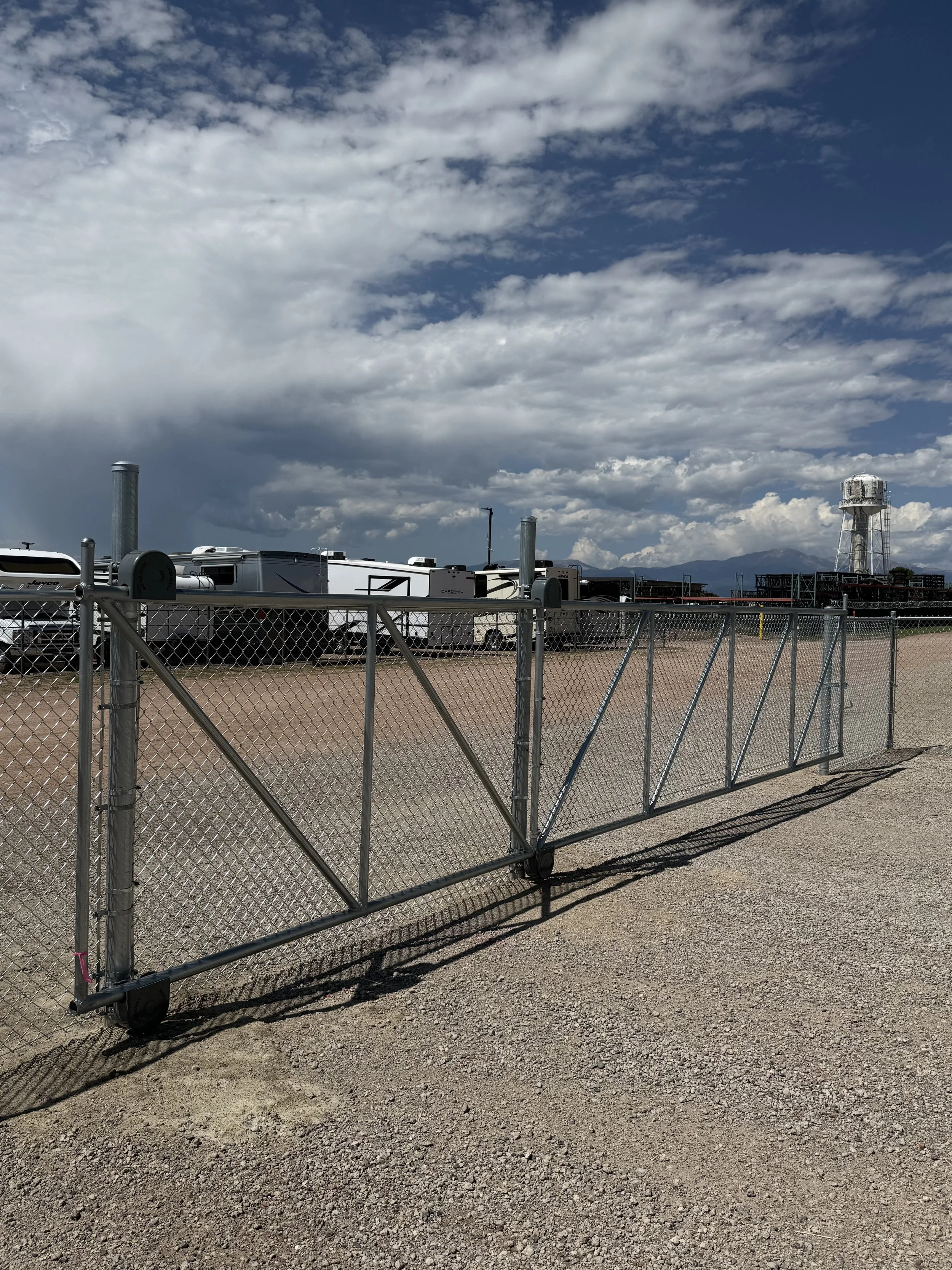 A chain-link fence gate in a dry, gravel lot with RVs and a water tower in the background under a cloudy sky.
