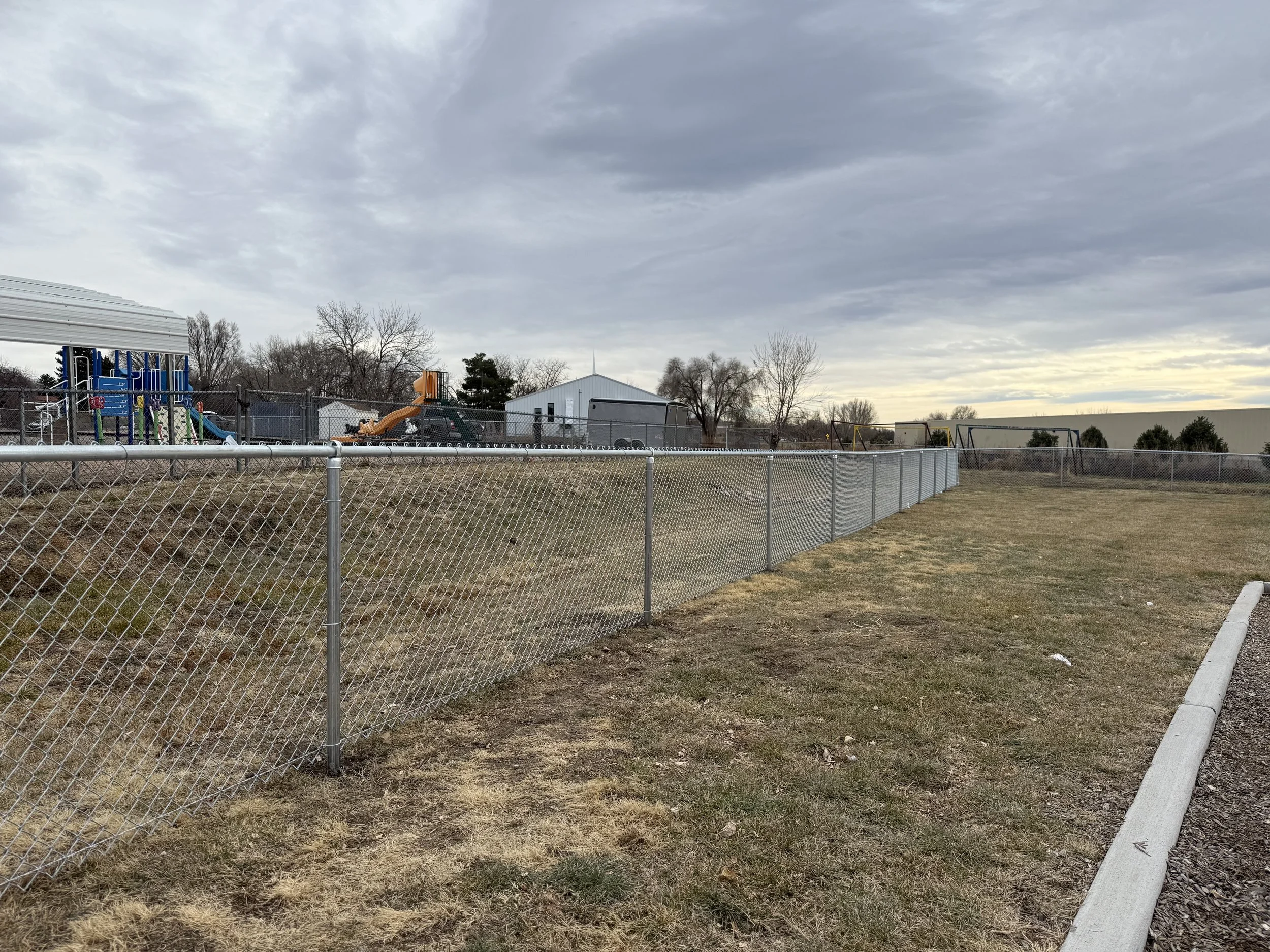 A fenced outdoor area with a playground and storage buildings under a cloudy sky.