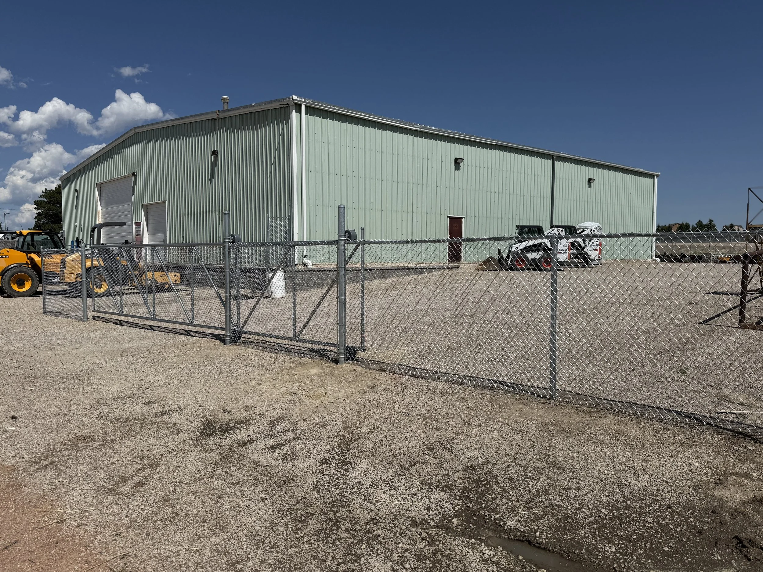 Large green metal industrial building with a chain-link fence in front, gravel ground, shipping doors, and vehicles including a small skid steer loader and skid steers in the background under a partly cloudy sky.